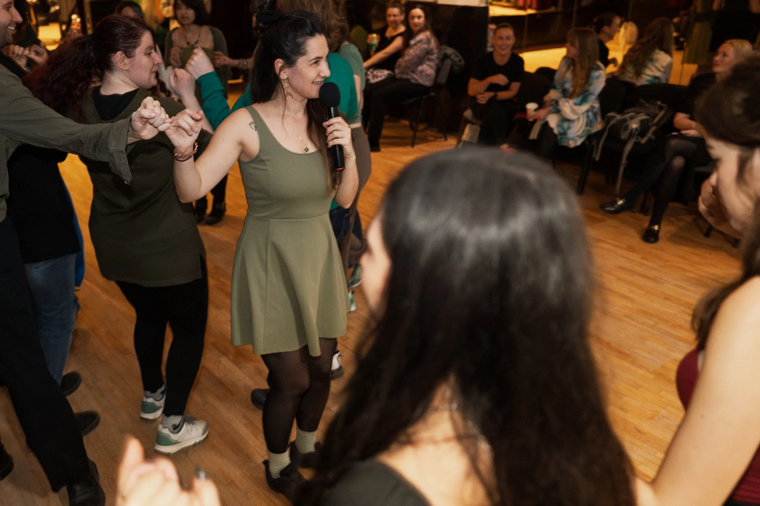 A woman holding a microphone in a green dress appears to be singing or speaking, surrounded by people in a social setting with wooden flooring and warm lighting.