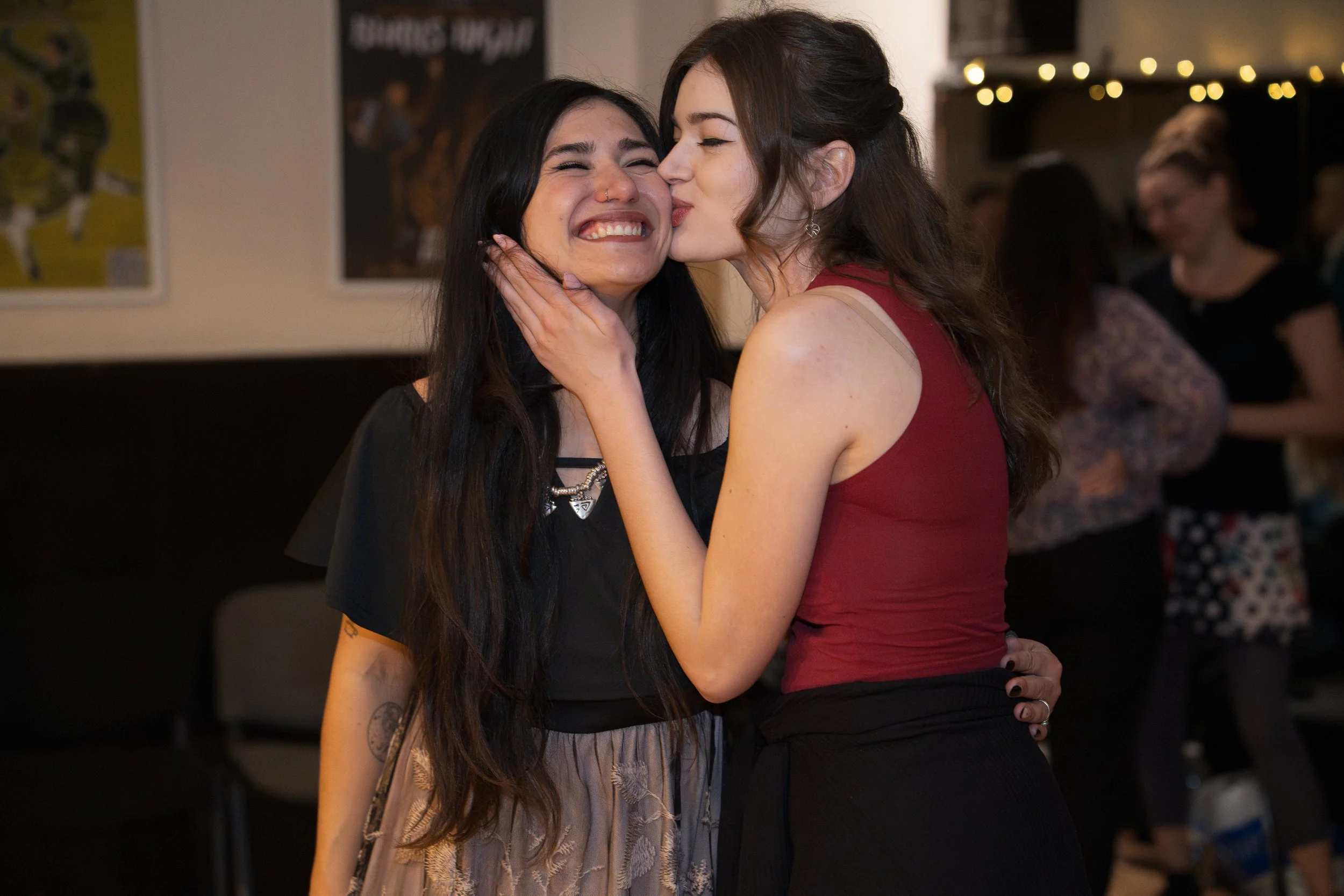 Two women embracing and smiling at each other at a social gathering, with other people in the background.
