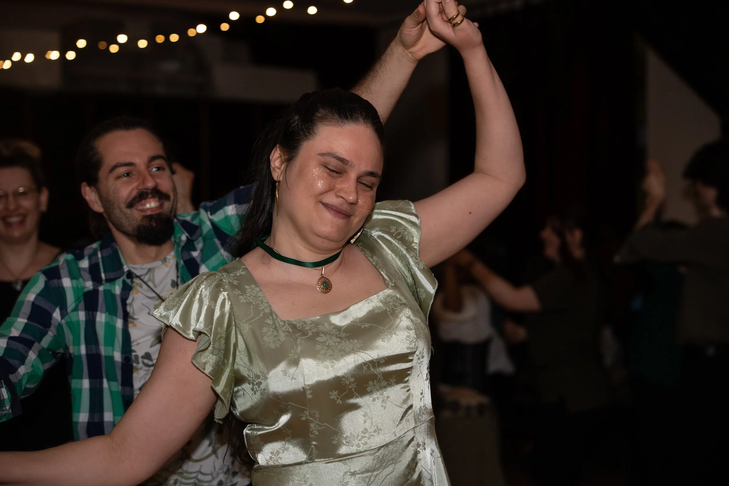 A woman with dark hair, wearing a shiny, patterned, light green dress, dancing with her arms raised and eyes closed, smiling. A man in a plaid shirt with facial hair and a big smile is dancing behind her. Other people are visible in the background at