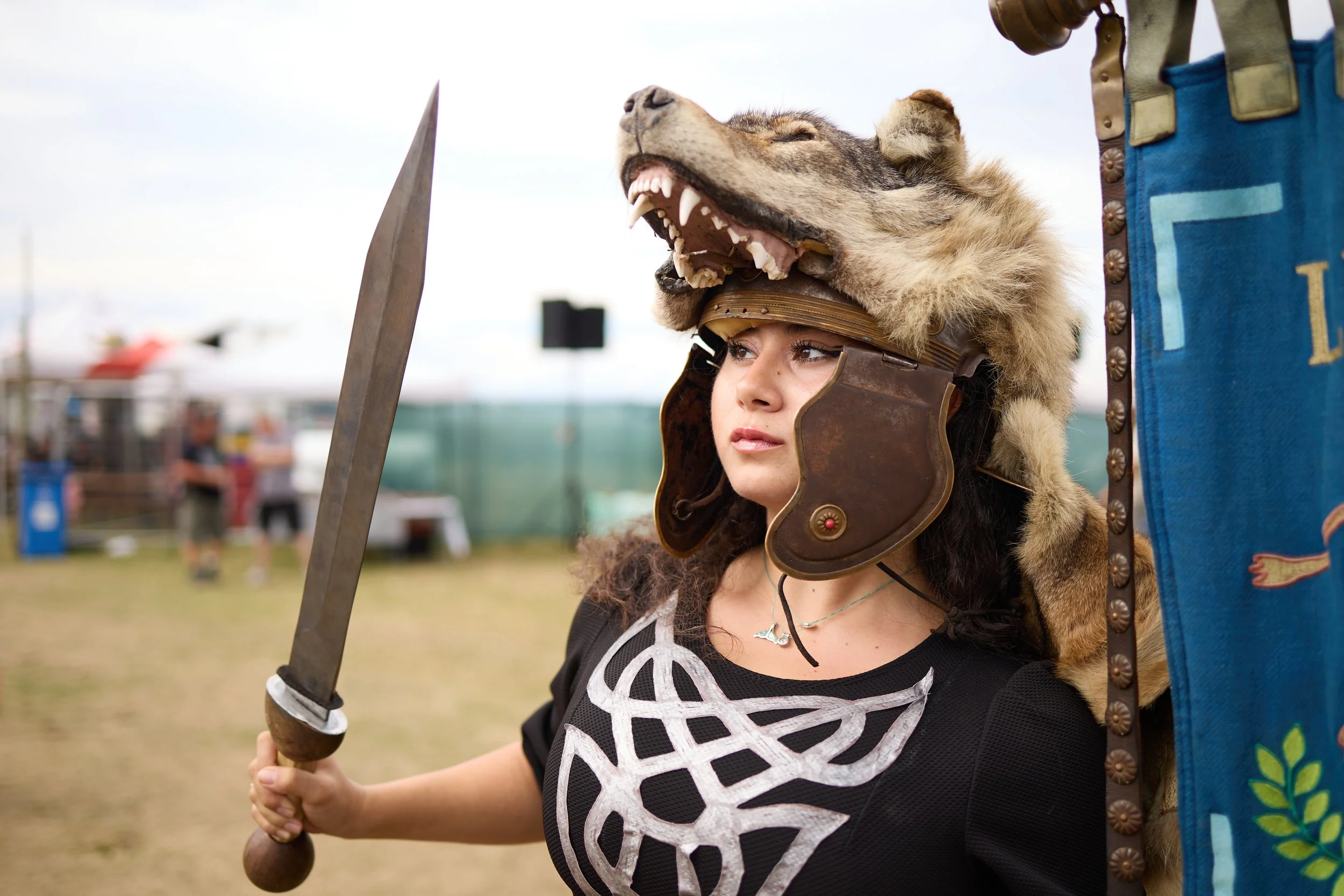 A young woman at an outdoor event dressed as a warrior, wearing a wolf's head helmet and holding a sword.