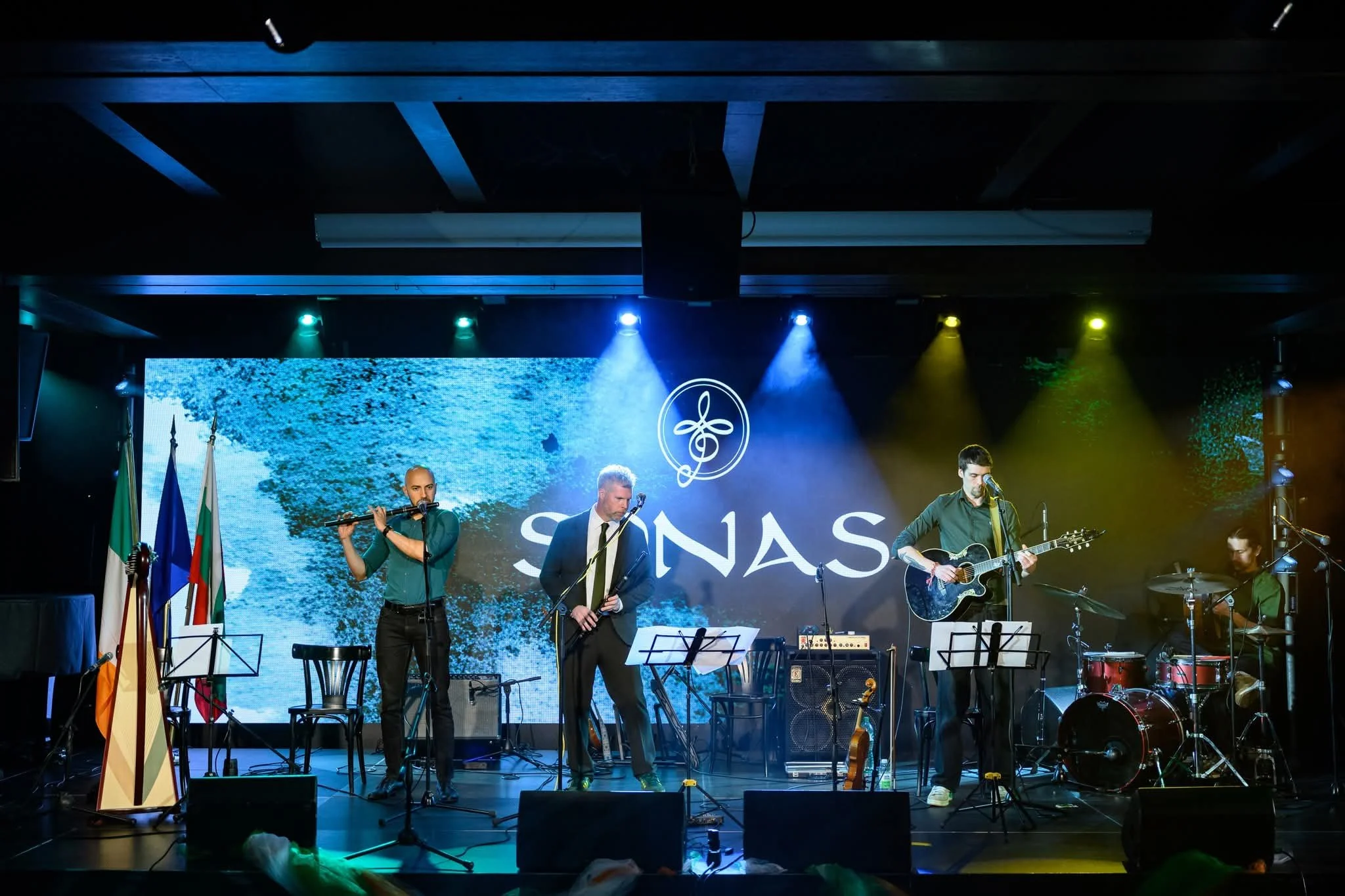 A band performs on stage with three musicians playing instruments and singing, a large screen displaying water behind them, and flags standing on the left side.