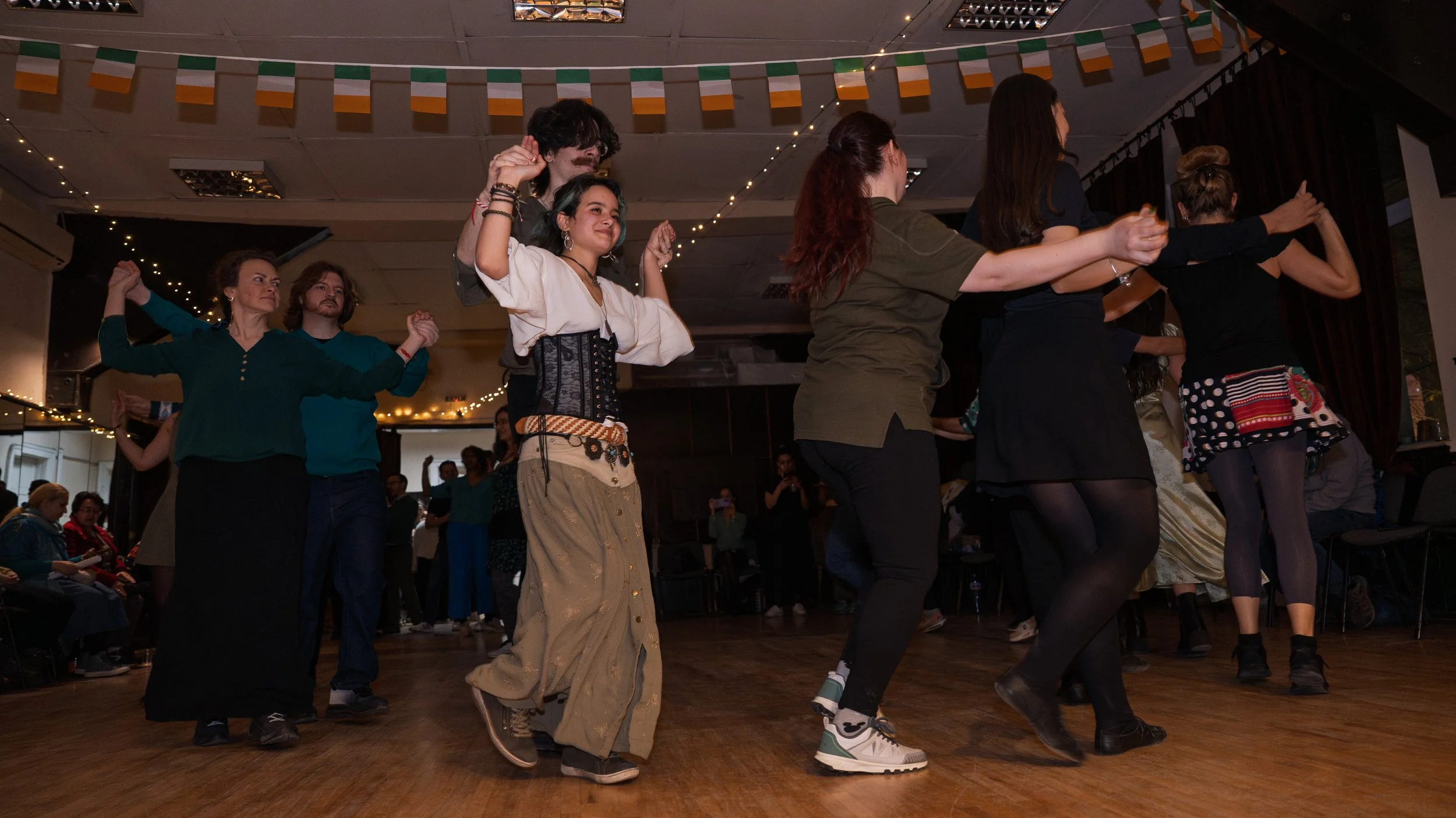 Group of people dancing together indoors, decorated with hanging Irish flags and string lights.