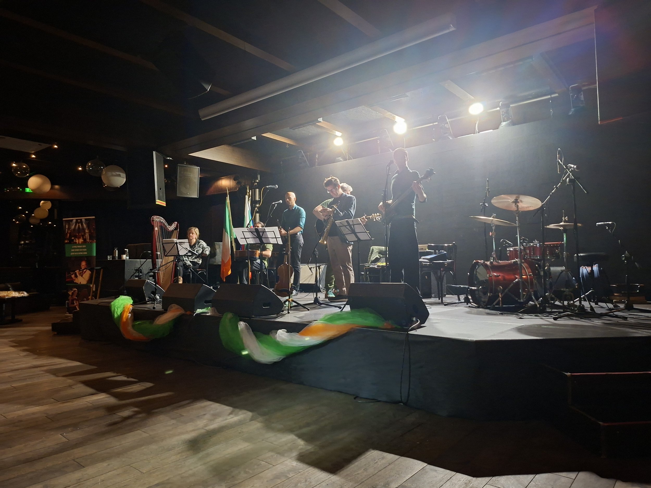 A stage with musicians performing in a dimly lit venue, with bright stage lights overhead and colorful decorations on the floor.