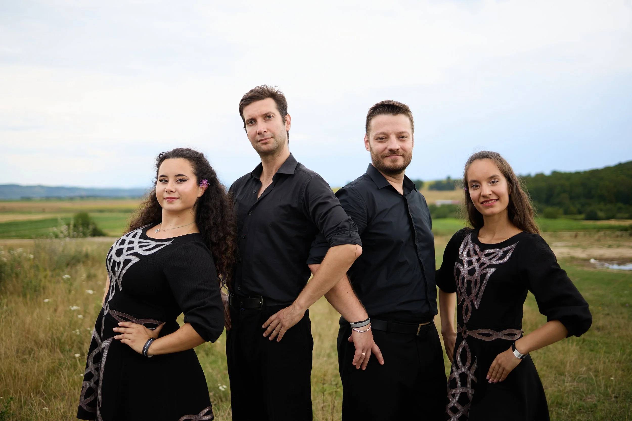 Four people, two women and two men, stand outdoors in a field with green trees and hills in the background. They are dressed in black clothing with patterned designs and pose confidently, looking at the camera.