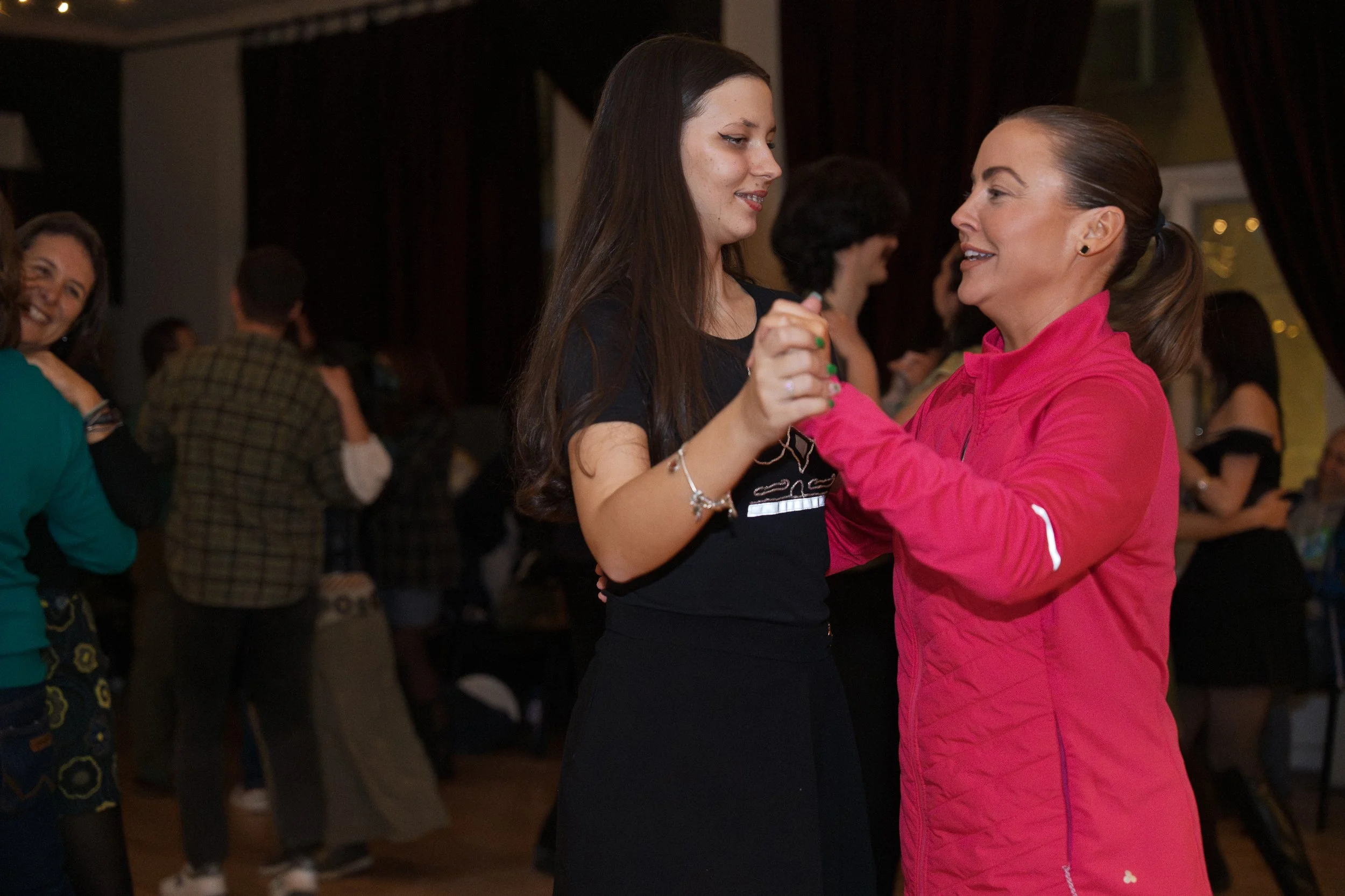 Two women dance together at a social gathering, smiling at each other, with other people in the background.