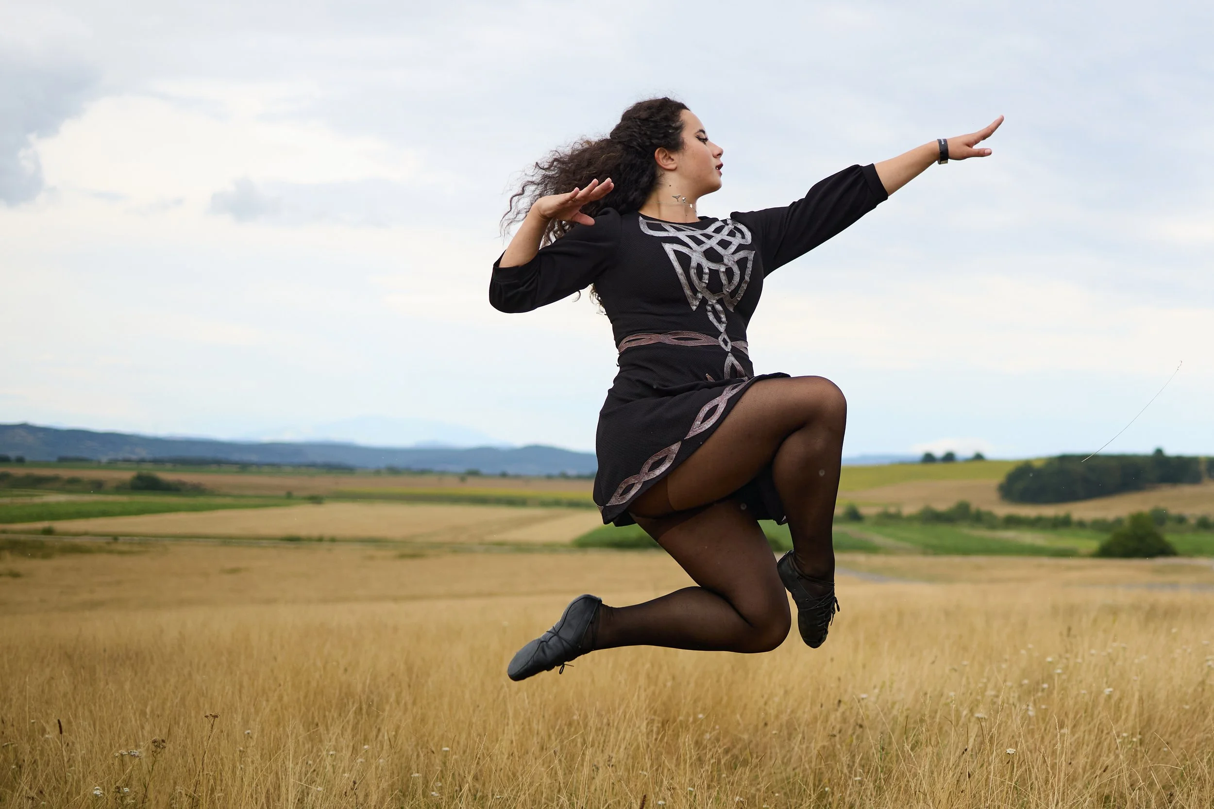 A woman in a black dress with silver patterns is seen jumping in a field of tall grass, with rolling hills and a cloudy sky in the background.
