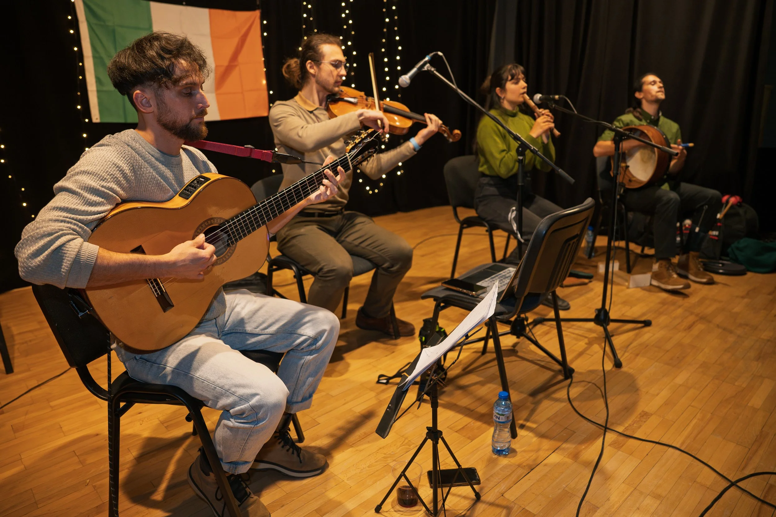 Four musicians performing on stage, playing instruments including guitar, violin, flute, and mandolin, with an Irish flag in the background and string lights decorating the stage.