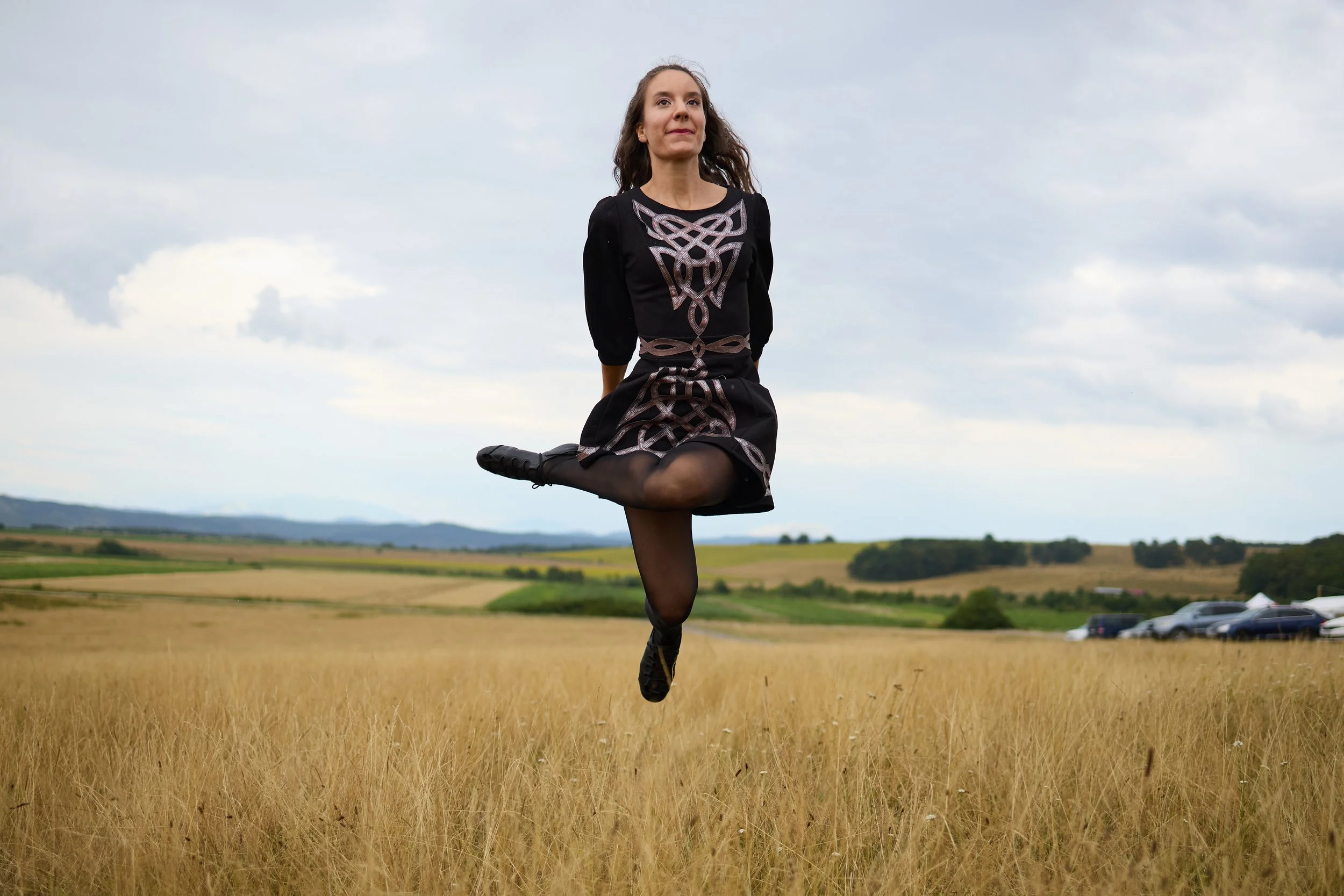 A woman in a black dress with intricate silver designs is standing on one leg in a field, with hills and a cloudy sky in the background.
