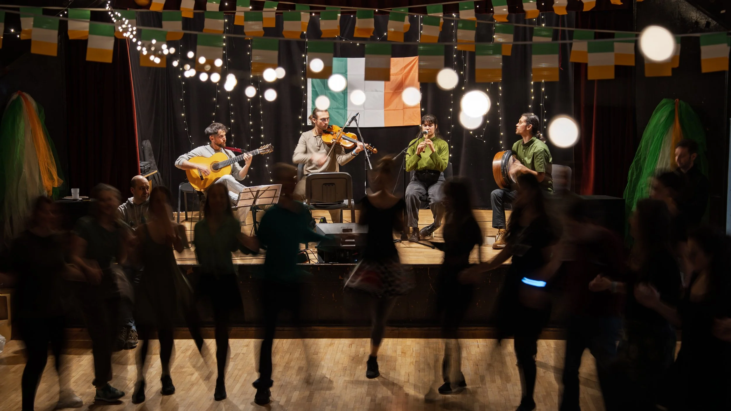 A musical performance on stage with four musicians playing instruments, Irish flags hanging above, and Irish flags in the background. Audience members are dancing and enjoying the event.