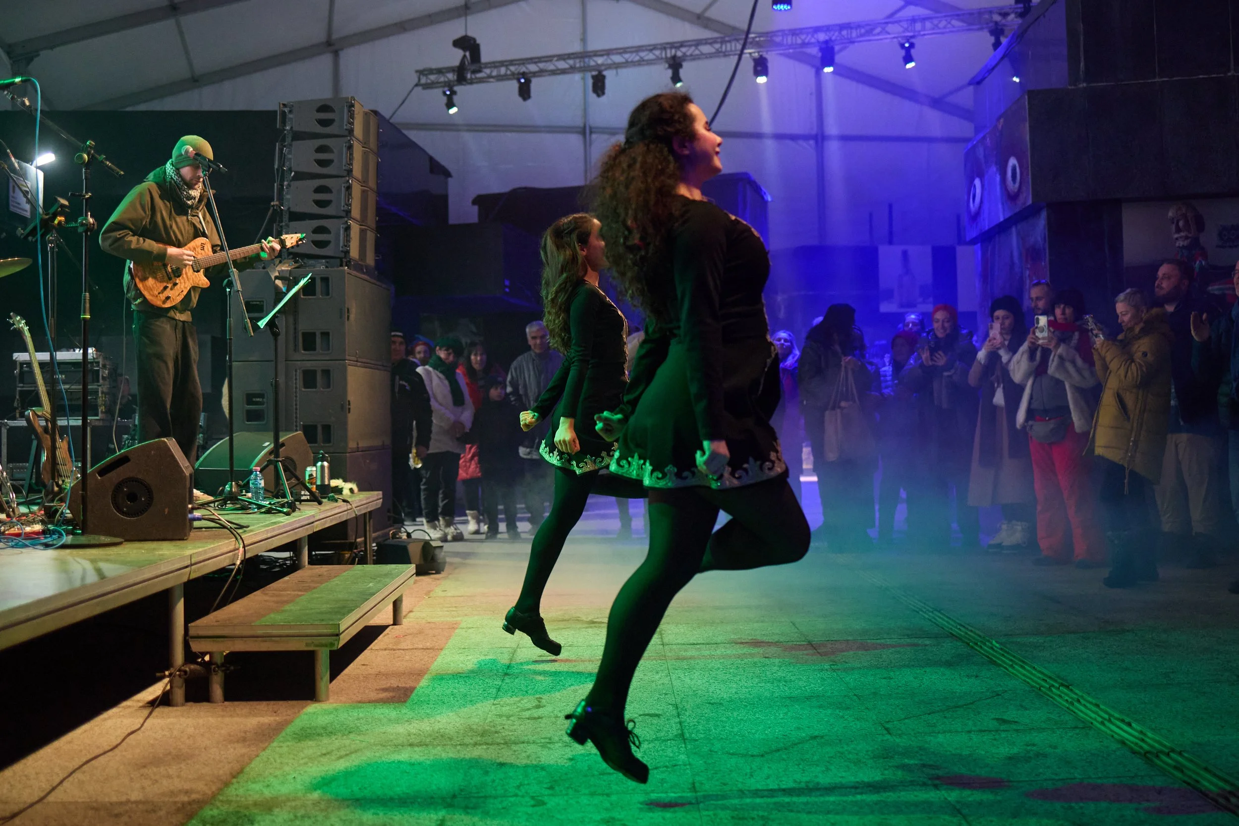 People dancing in a dimly lit indoor venue with a band performing on stage, including a guitarist, and the audience watching.