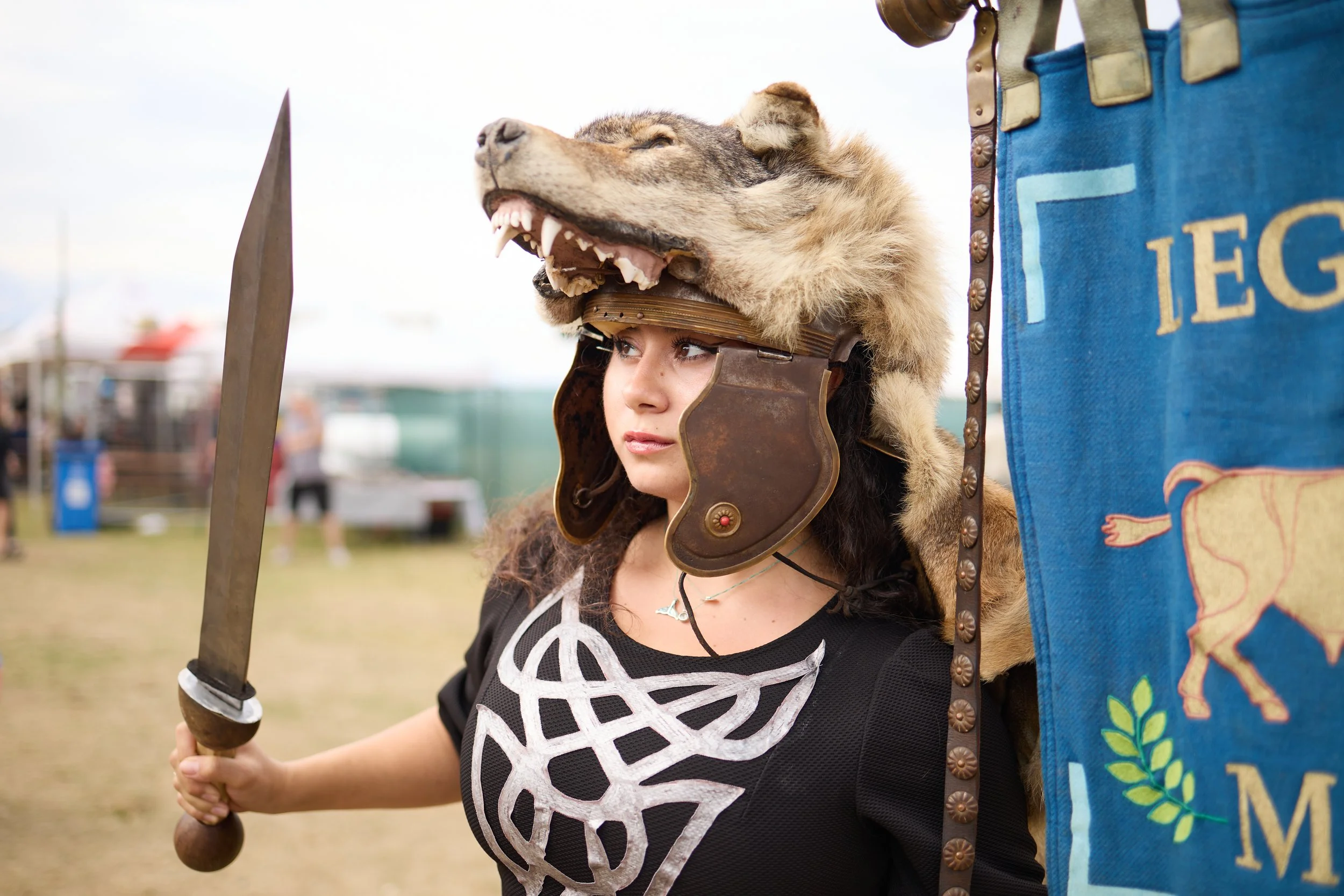 A woman wearing a helmet with a lion's head on top, holding a staff, at an outdoor event.