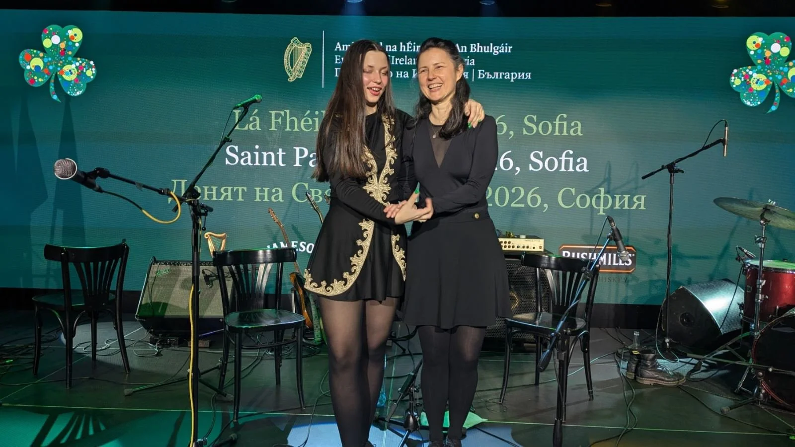 Two women standing on stage, smiling, with musical instruments and microphones around them. Background displays text in multiple languages and graphics of clovers.