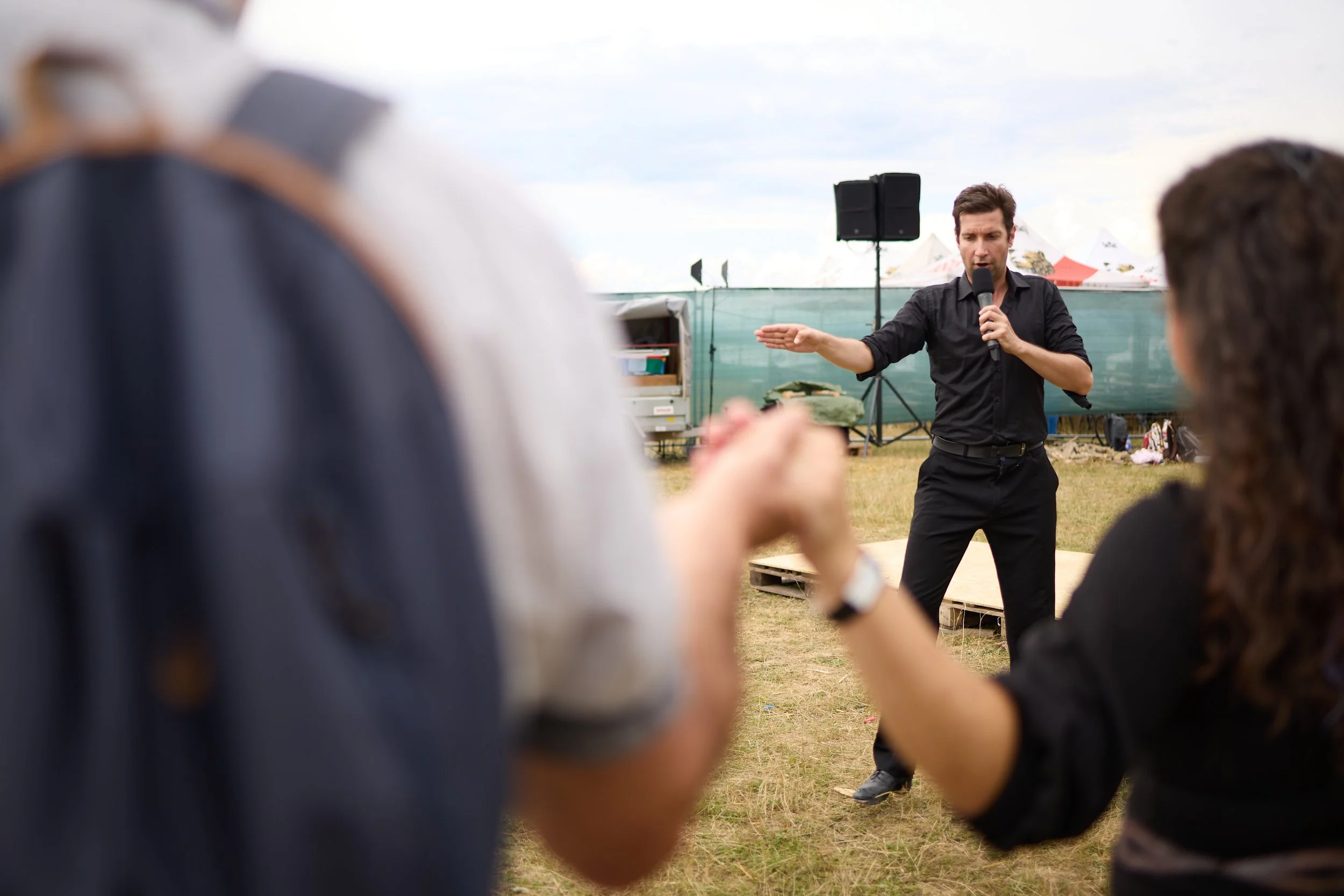 A man in black clothes delivering a speech with a microphone at an outdoor event, with tents and speakers in the background. Several audience members are in the foreground.