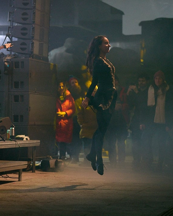 Woman in black outfit jumping in front of an audience at an event or concert.