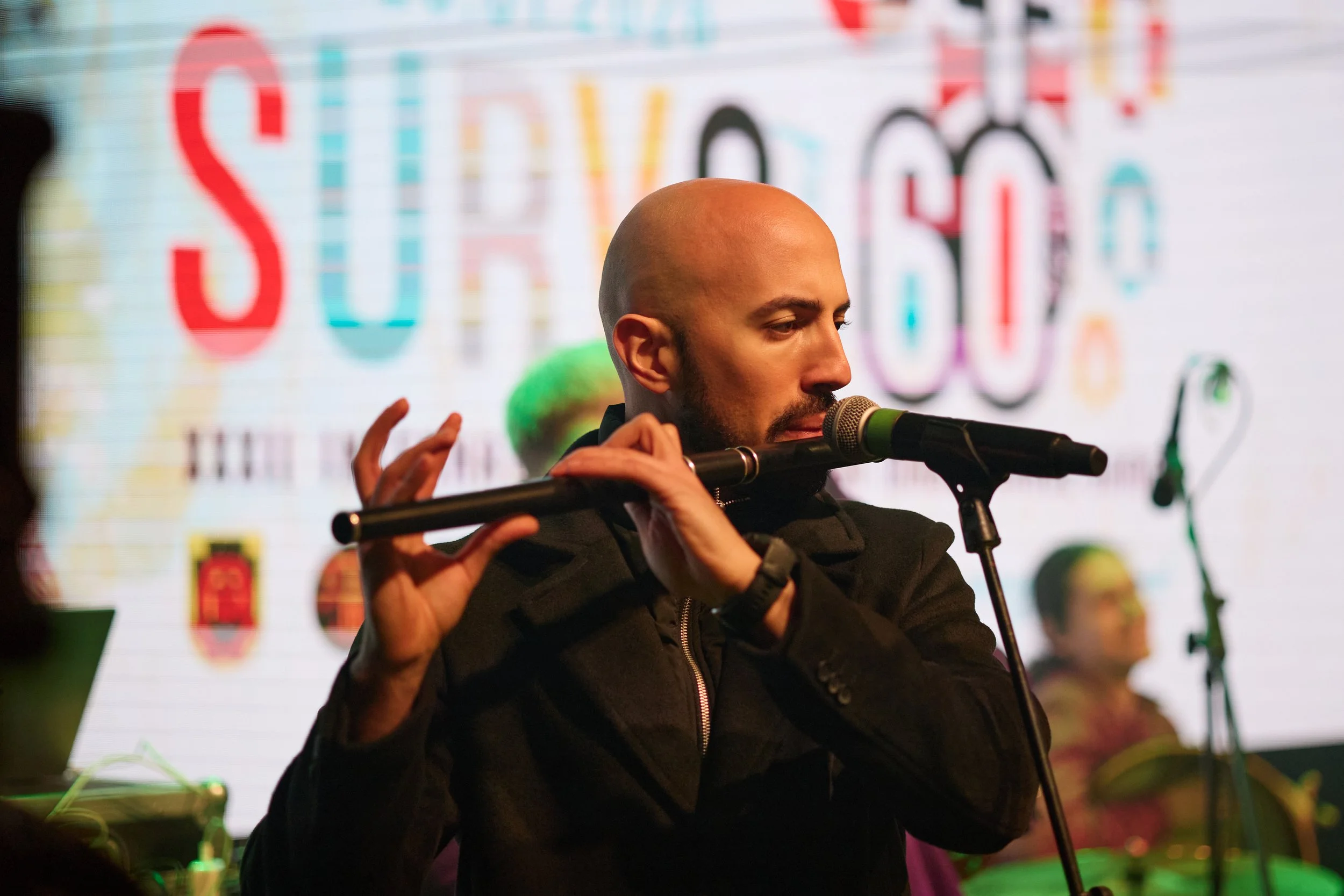 A man with a shaved head and beard playing a flute at a music event.