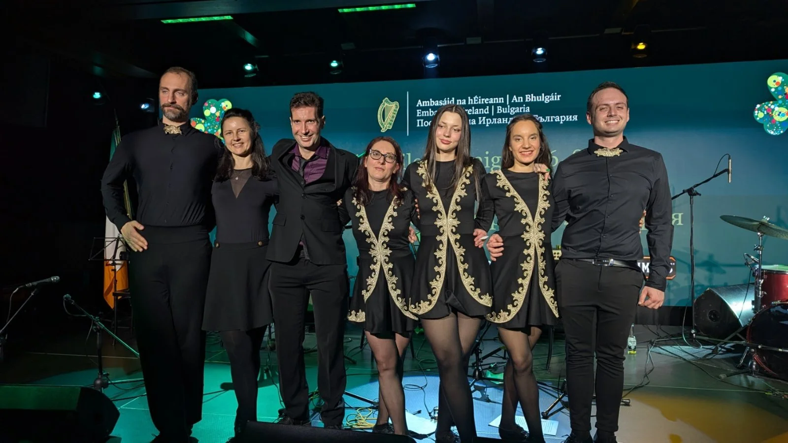 Group of seven performers on stage, dressed in black with gold embroidery, posing for photo after performance, with musical instruments and a green backdrop behind them.