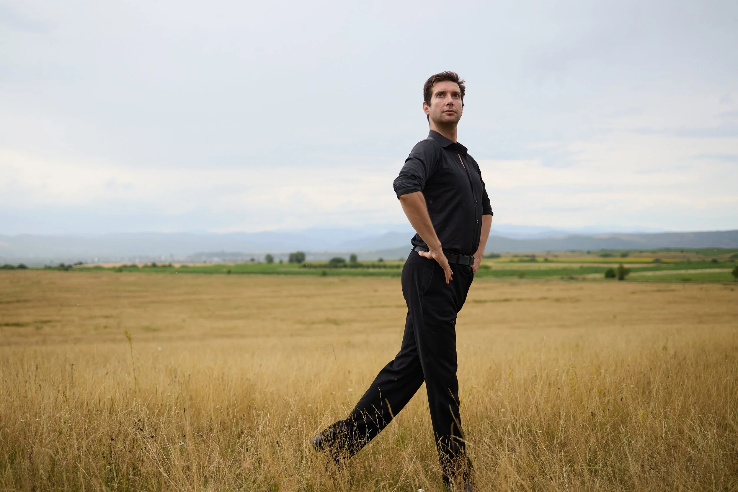 A man in black clothing walking through a field of tall grass with a cloudy sky and distant mountains in the background.