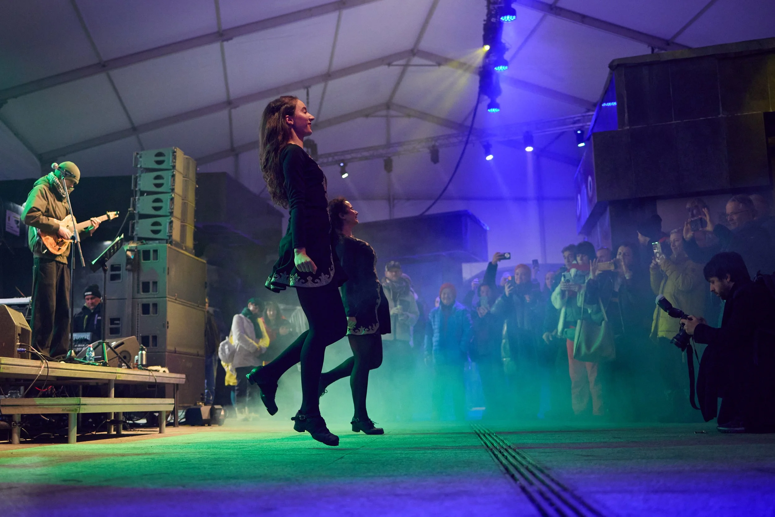 A woman and a woman dance on a stage during a performance inside a large tent with an audience watching.