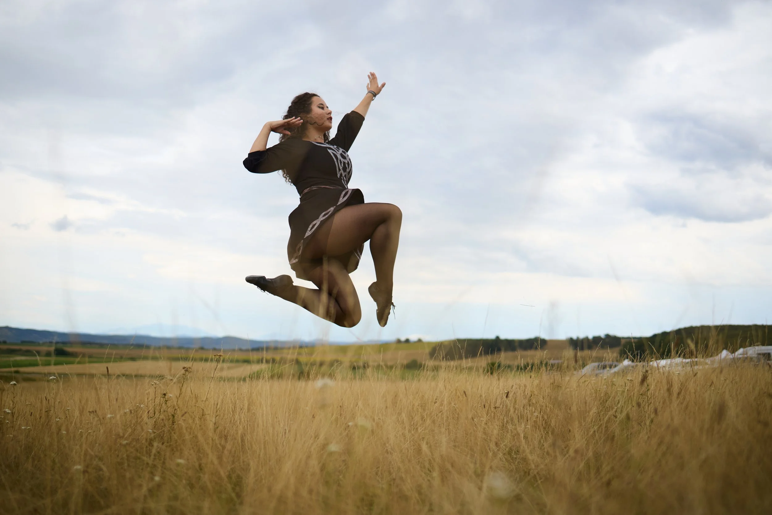 A woman in black dance attire is captured mid-air in a leap across a grassy field, with her arm raised and eyes closed against a cloudy sky and countryside landscape.