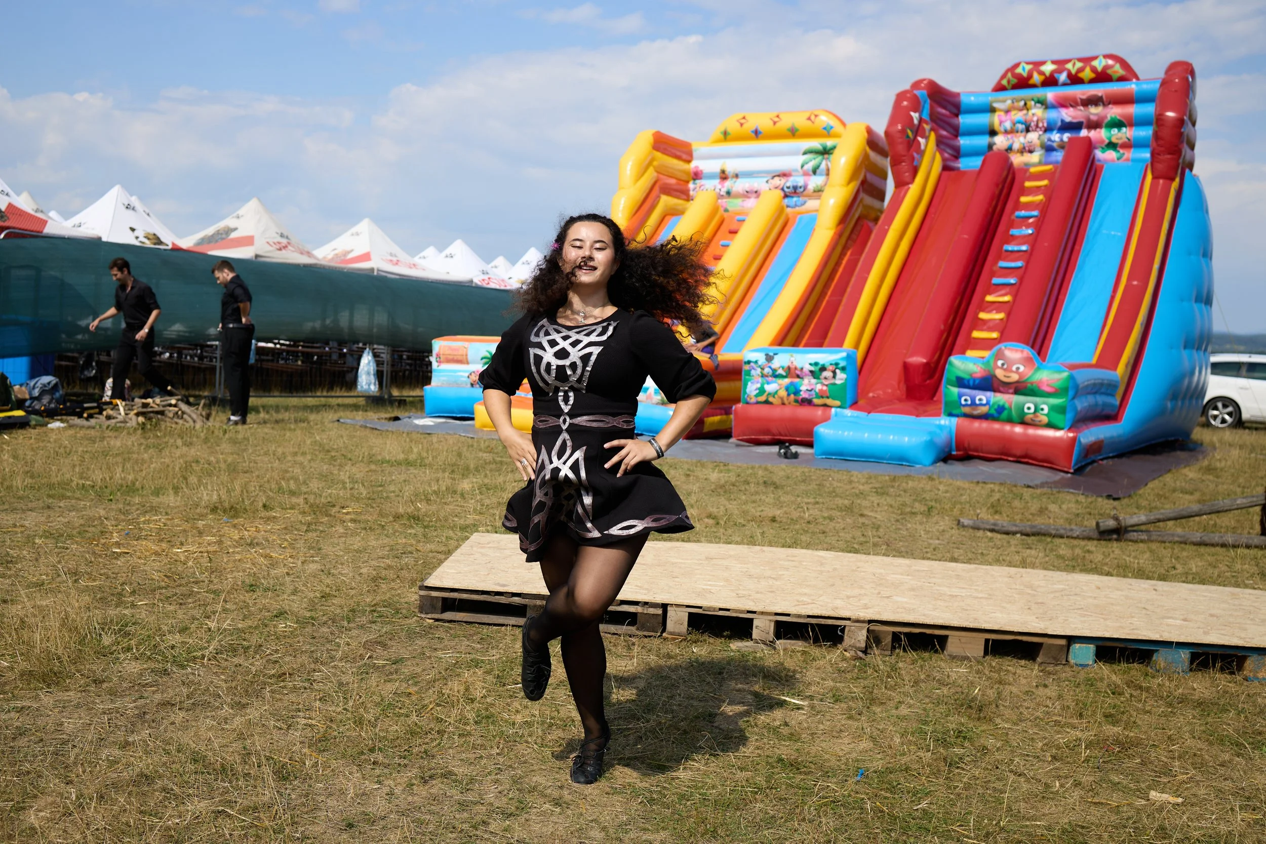 A woman with curly hair wearing a black dress smiling and running in front of inflatable slides at an outdoor fair or amusement park under a partly cloudy sky.