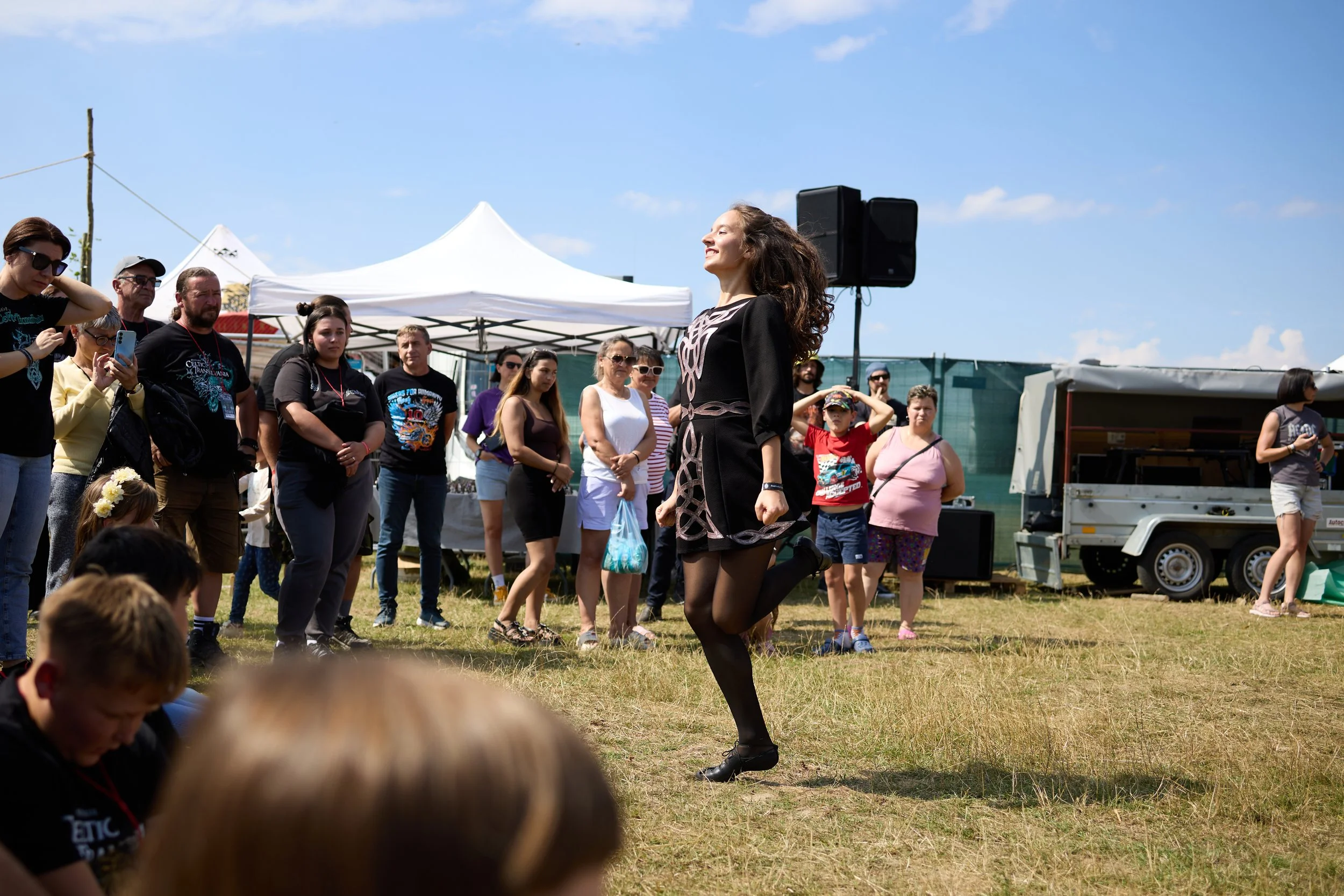 A woman dancing outdoors at a festival or event with a crowd of people watching. The sky is clear and blue, with tents and equipment in the background.