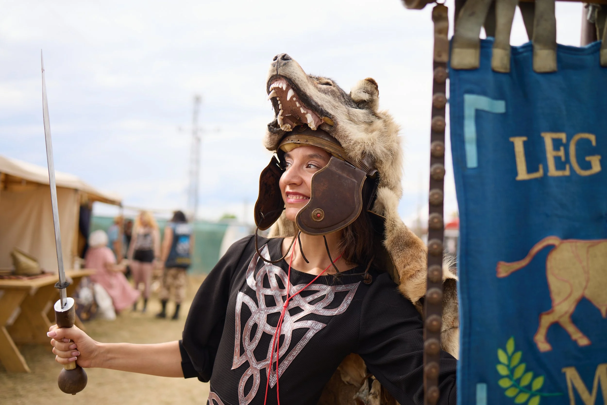 A woman smiling while wearing a helmet with a wolf's head on top at an outdoor event.