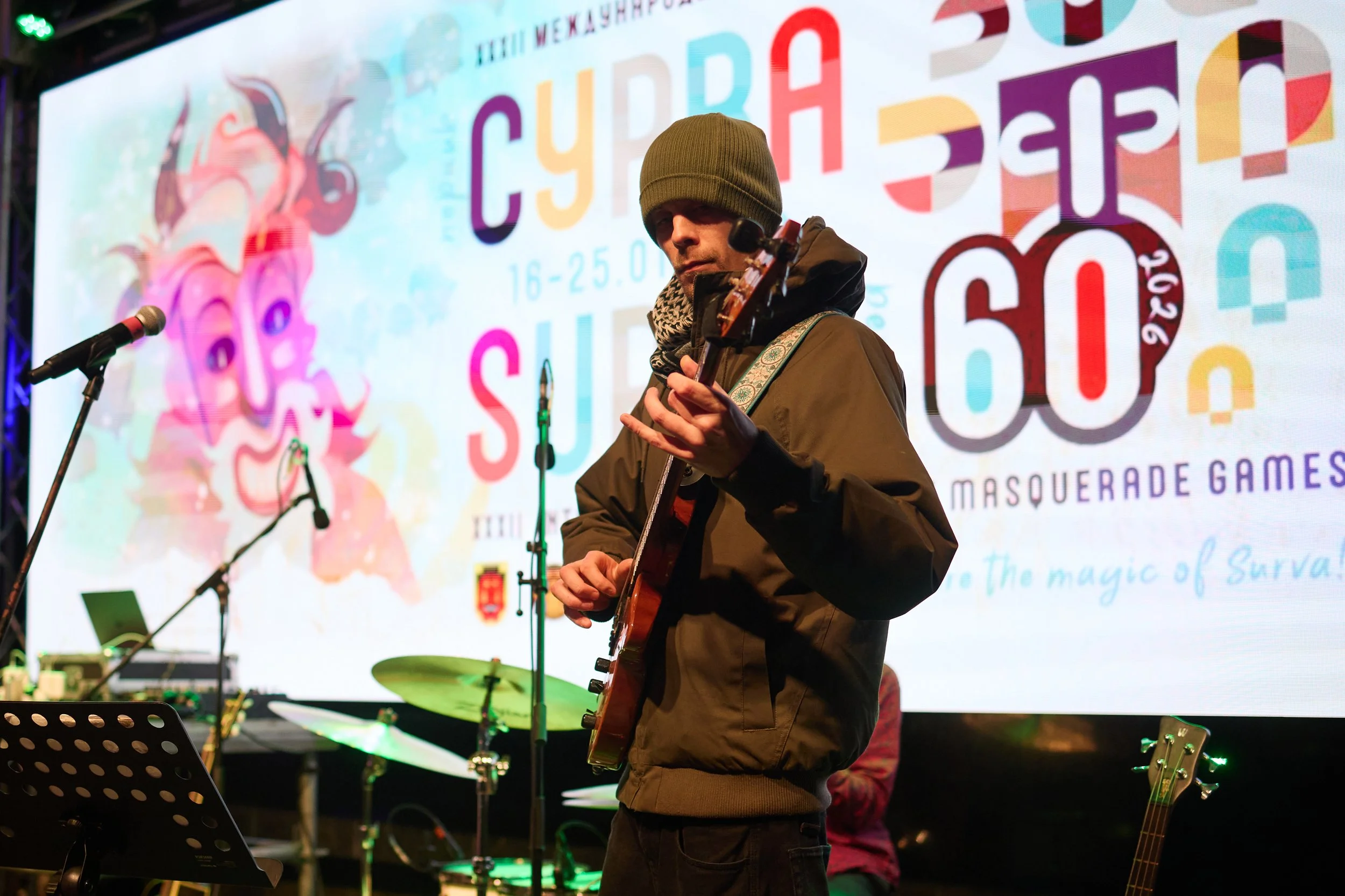 A man wearing a gray beanie and brown jacket playing an acoustic guitar on stage, with a colorful event banner in the background.