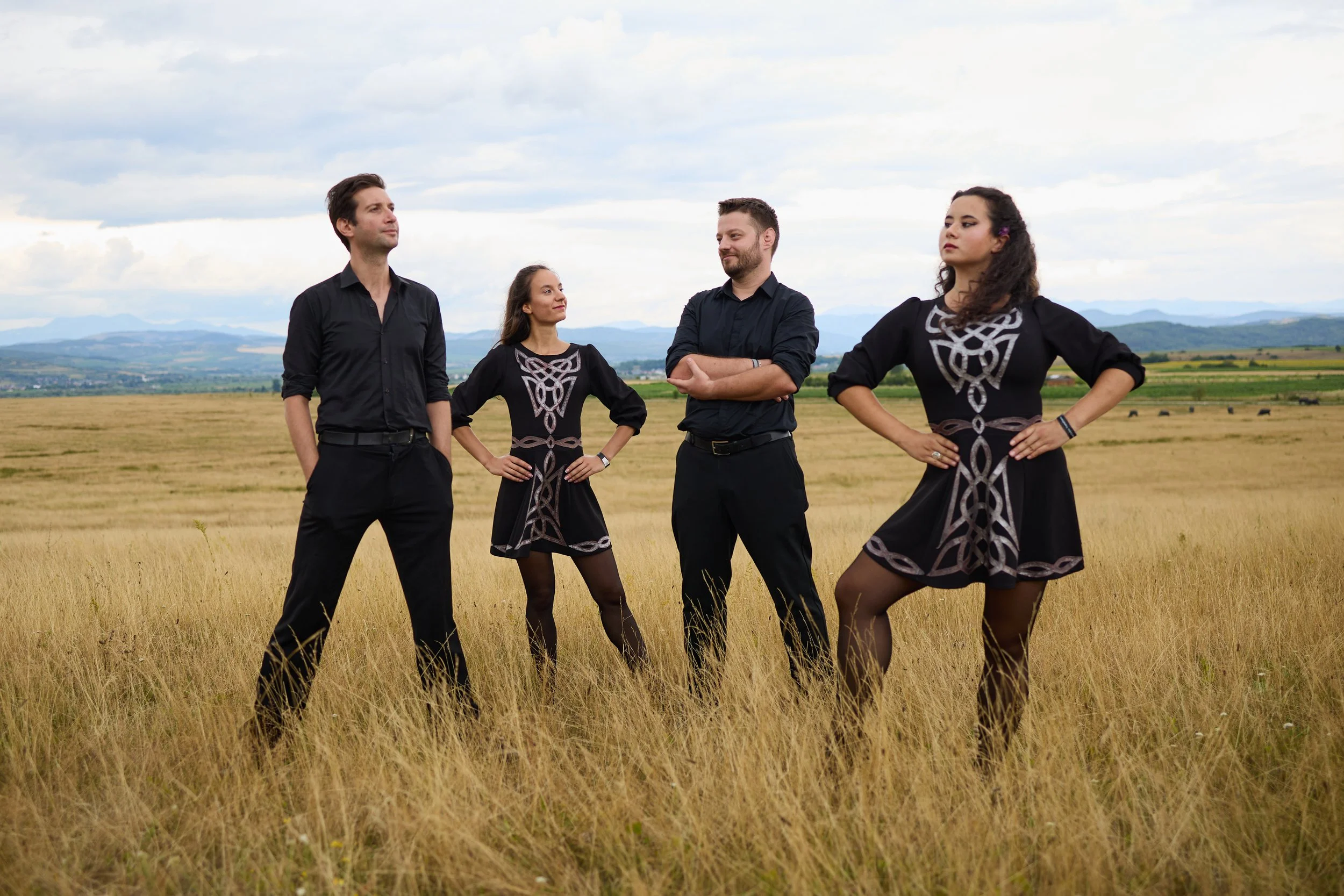 Four people in black clothing with Celtic designs on a grassy field under a cloudy sky.