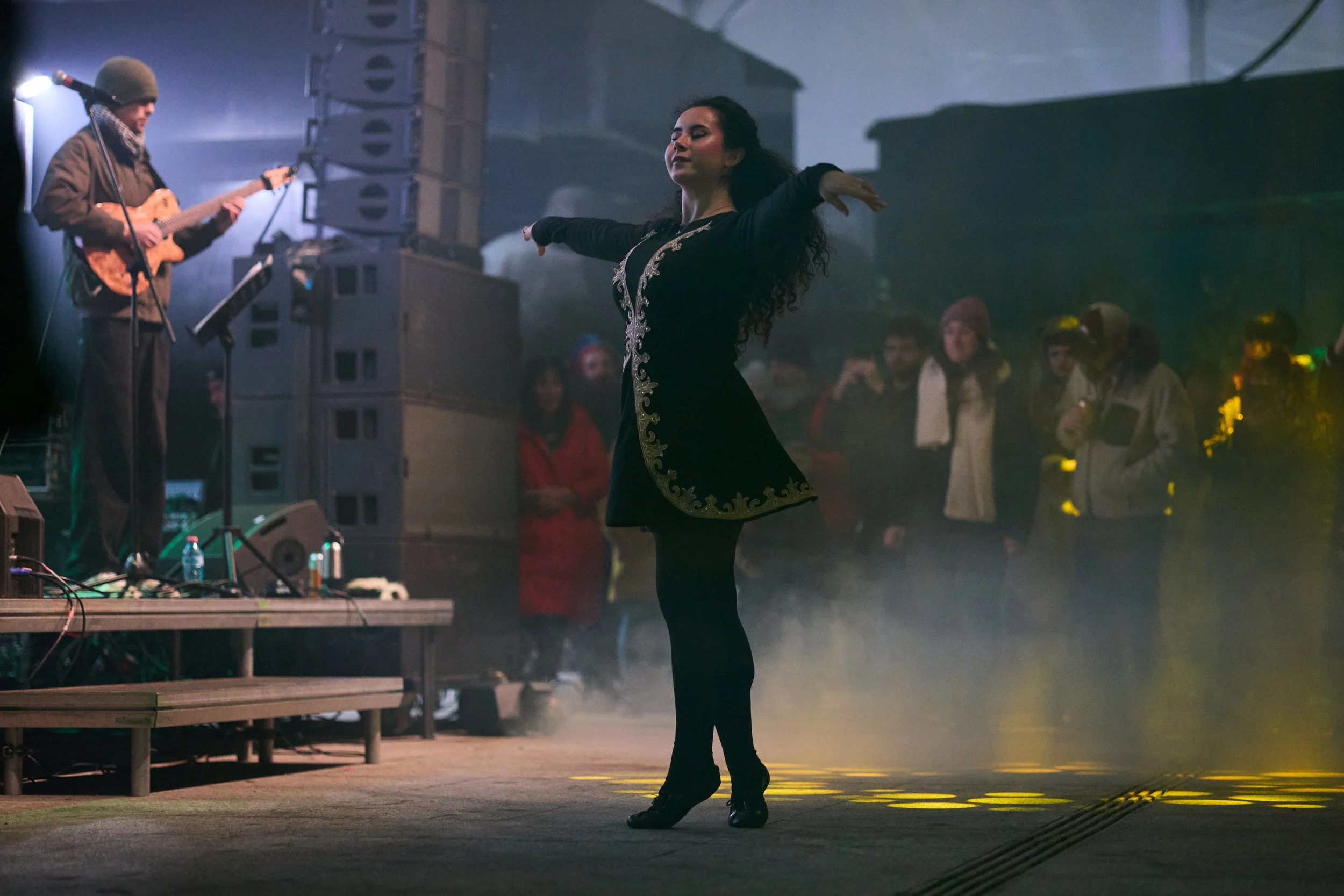 A woman in a black dress with decorative embroidery dances on a stage with her arms outstretched. A guitarist in a beanie and jacket plays beside her. Several audience members watch in the background, and stage equipment and speakers are visible.