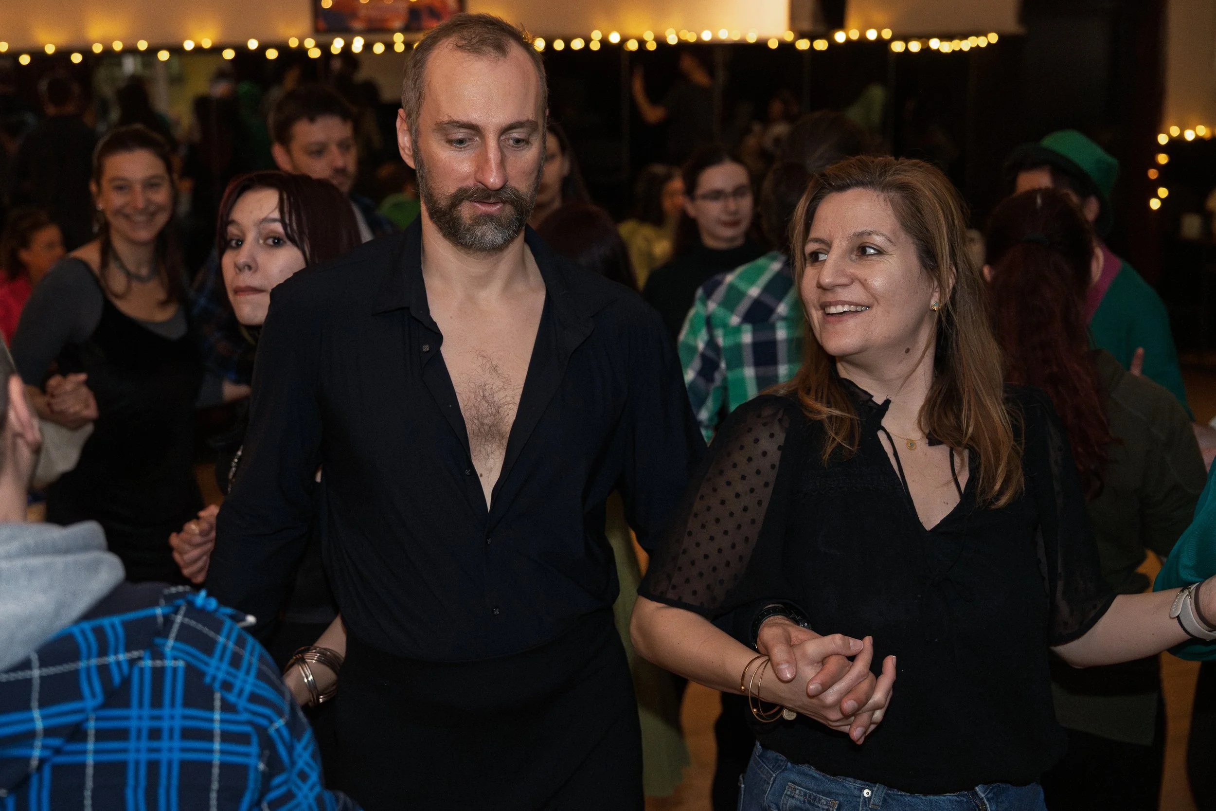 A group of people at a social gathering, with a man and woman in the foreground holding hands, smiling and talking. The background features warm lighting and a crowd of other guests.