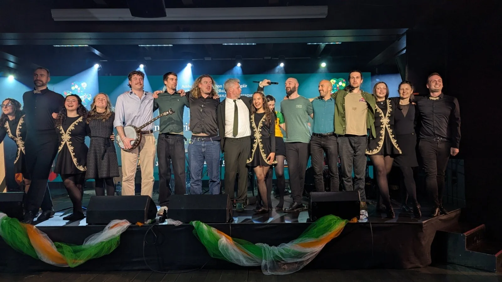 Group of people standing on stage after a performance or event, with some holding musical instruments and others embracing, smiling, or looking towards the audience.
