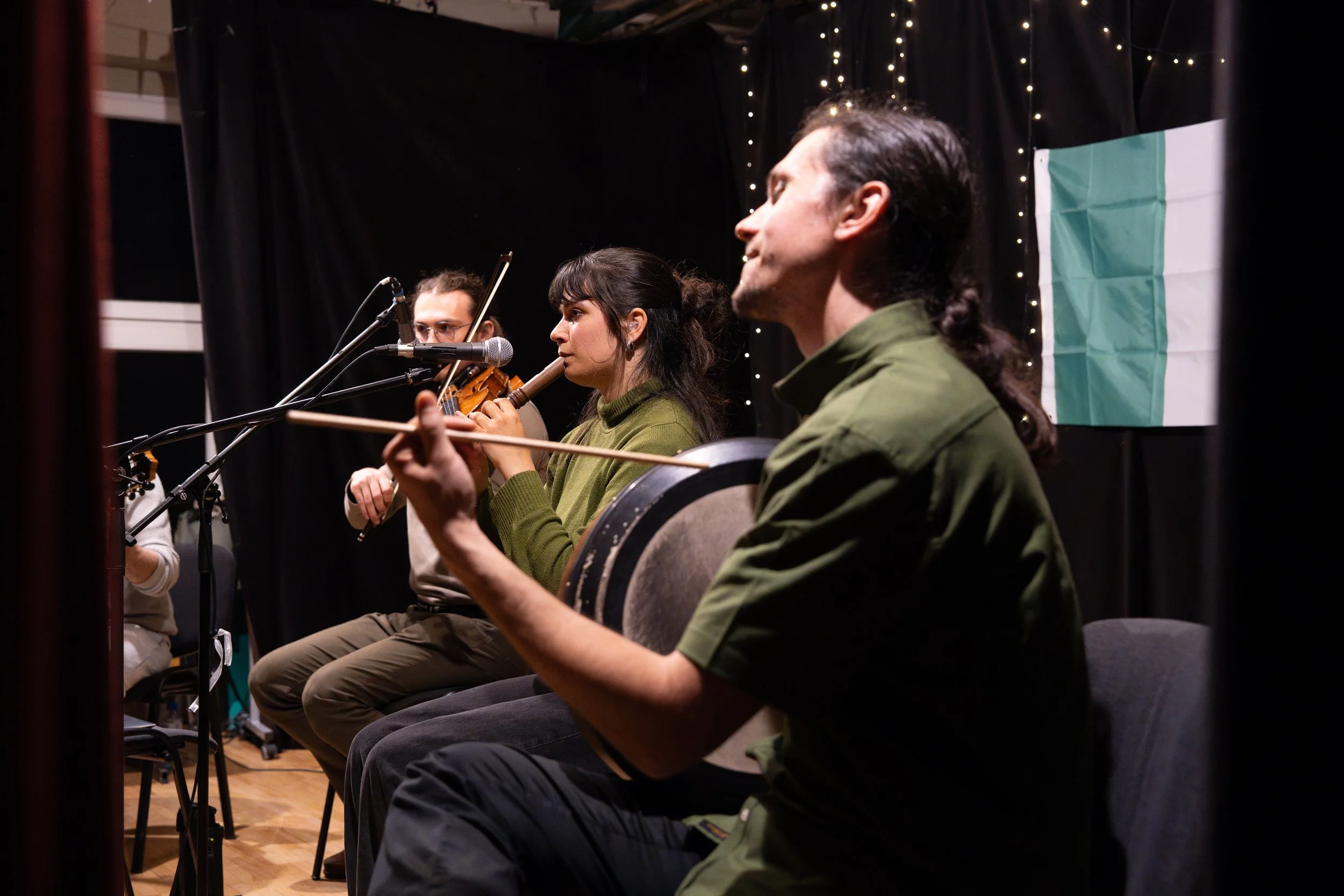 Three musicians seated in a row, playing instruments during a performance. The woman in the center is playing a flute, while the man on the right is playing a drum. The woman in the background appears to be playing a string instrument. They are on a 