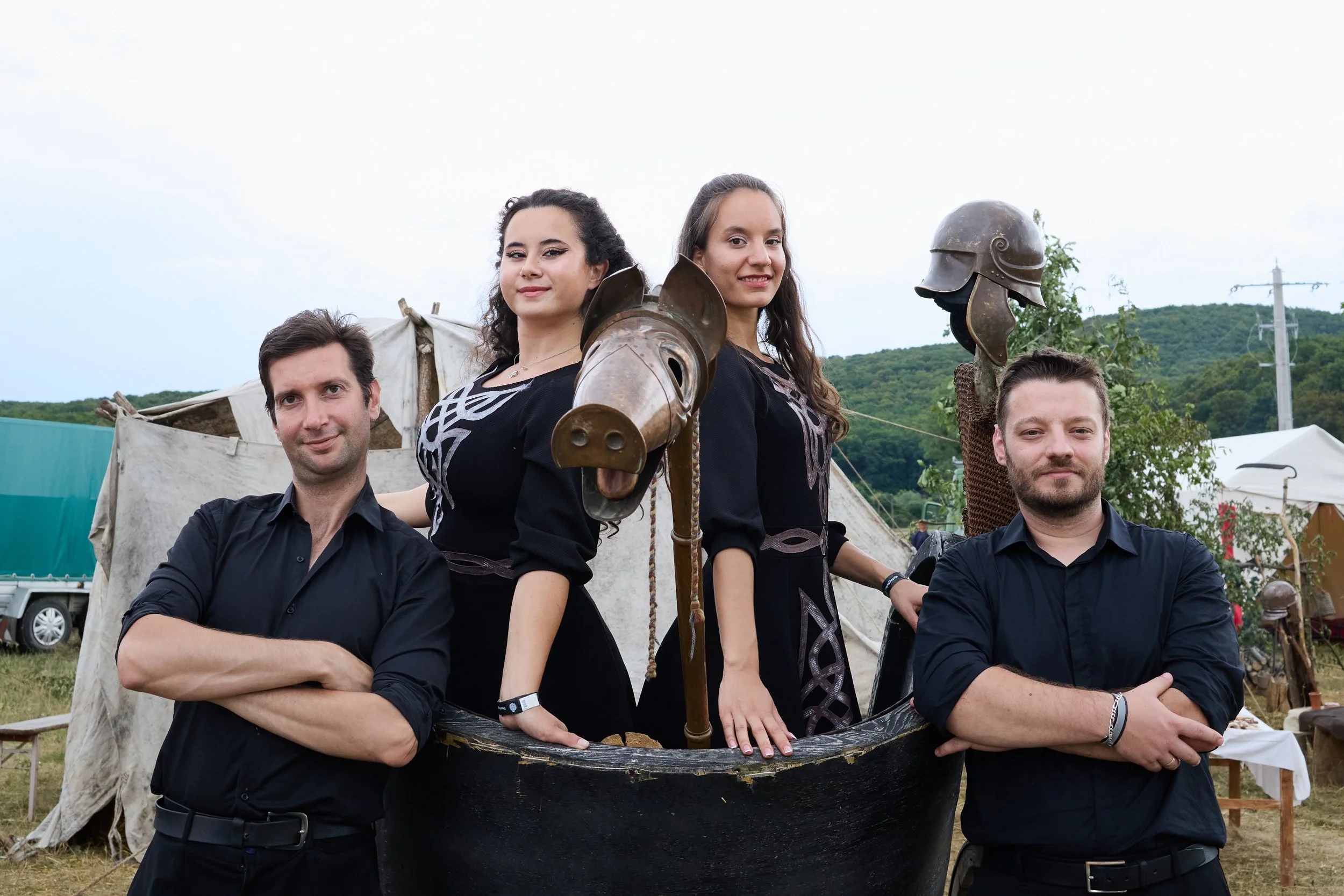 Four people, two men and two women, standing outdoors during daytime, with tents and greenery in the background. The women wear armor and hold shields, with one woman also holding a spear, suggesting they are reenactors or at a themed event.