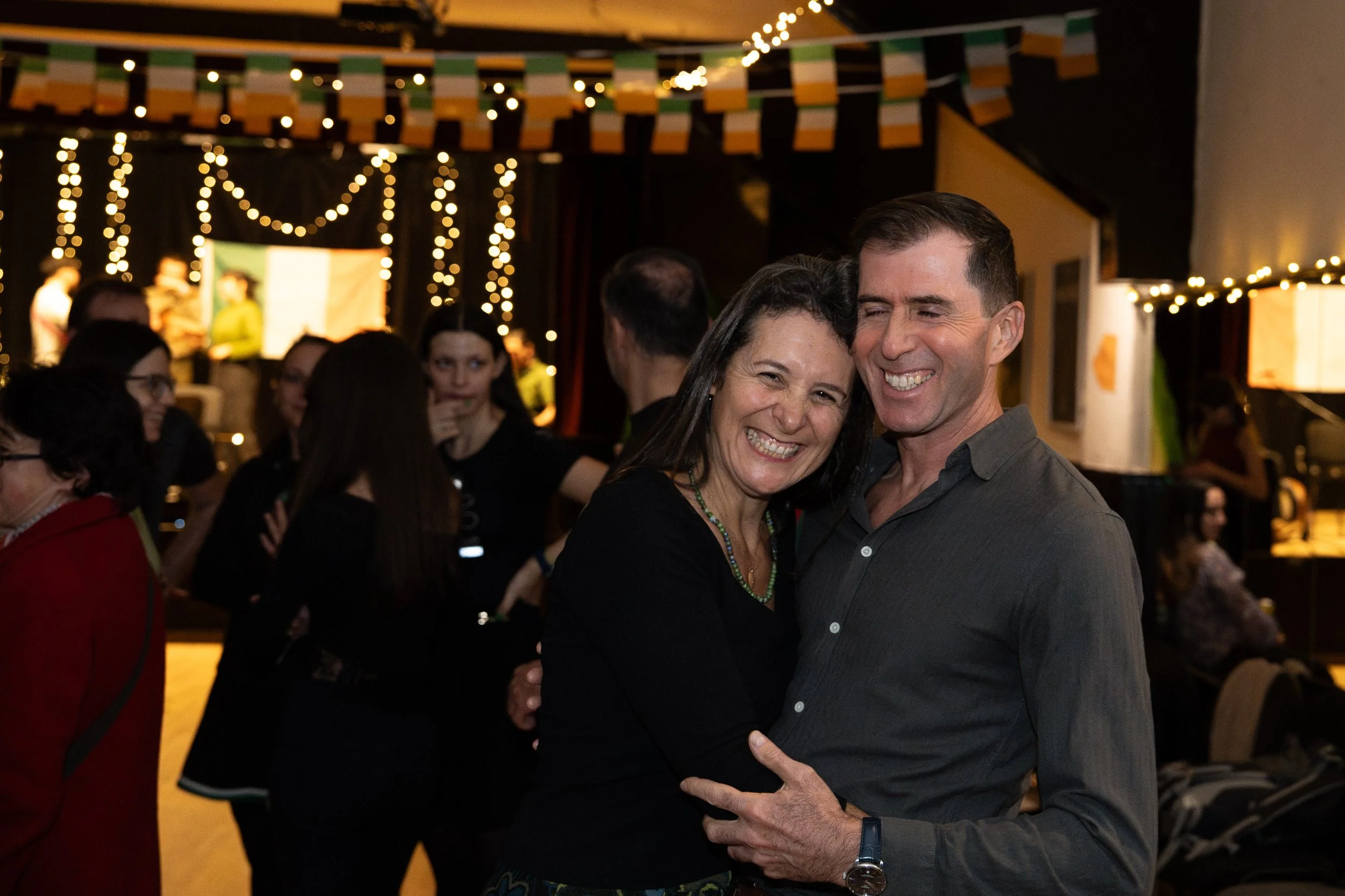 A couple is dancing closely and smiling at each other in a dimly lit room decorated with string lights and Irish flags, with other guests in the background.
