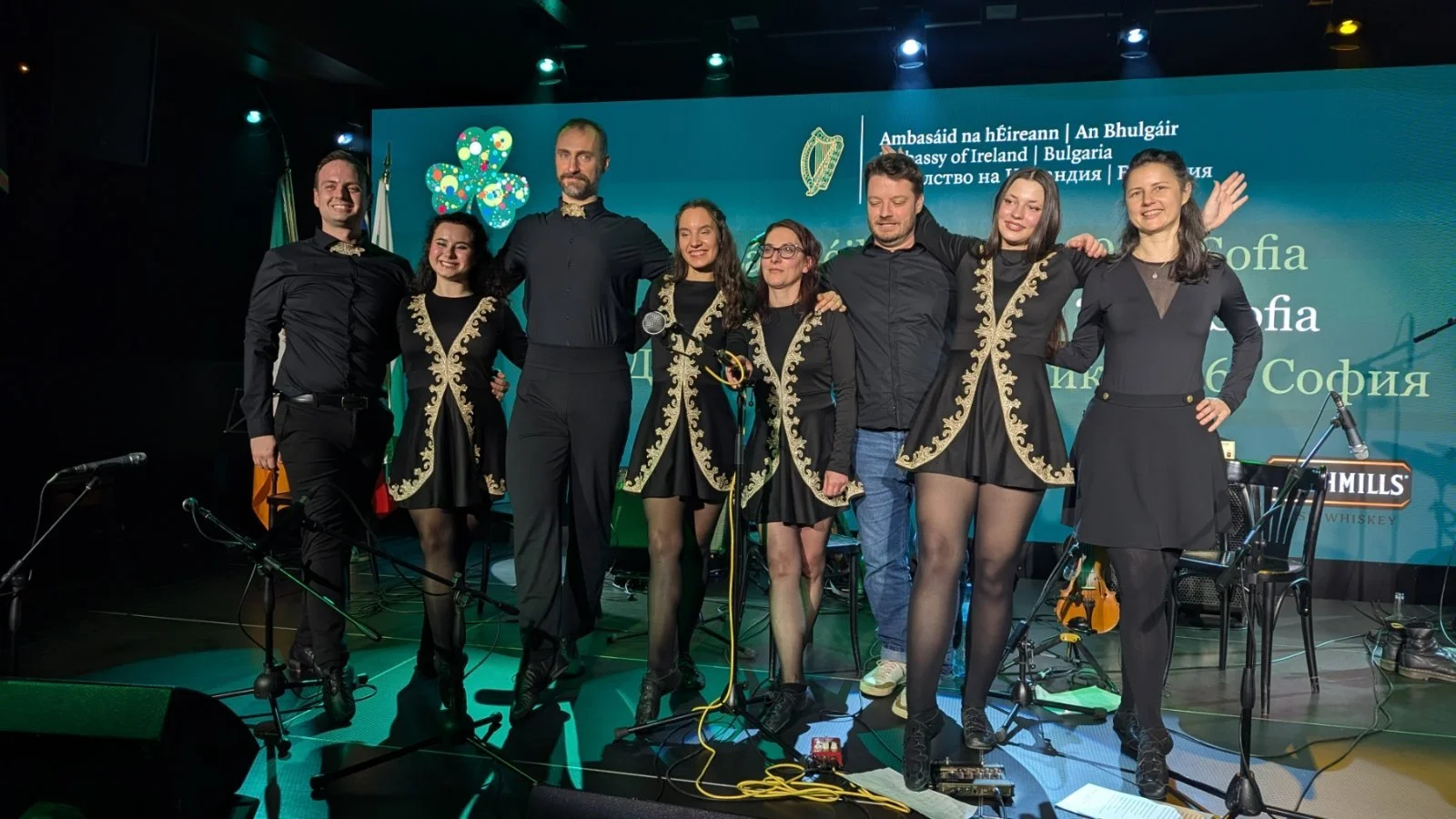 Group of people standing on stage at an event, dressed in coordinated black outfits with gold embroidery, smiling and posing for a photo, with a blue background displaying event details in multiple languages.
