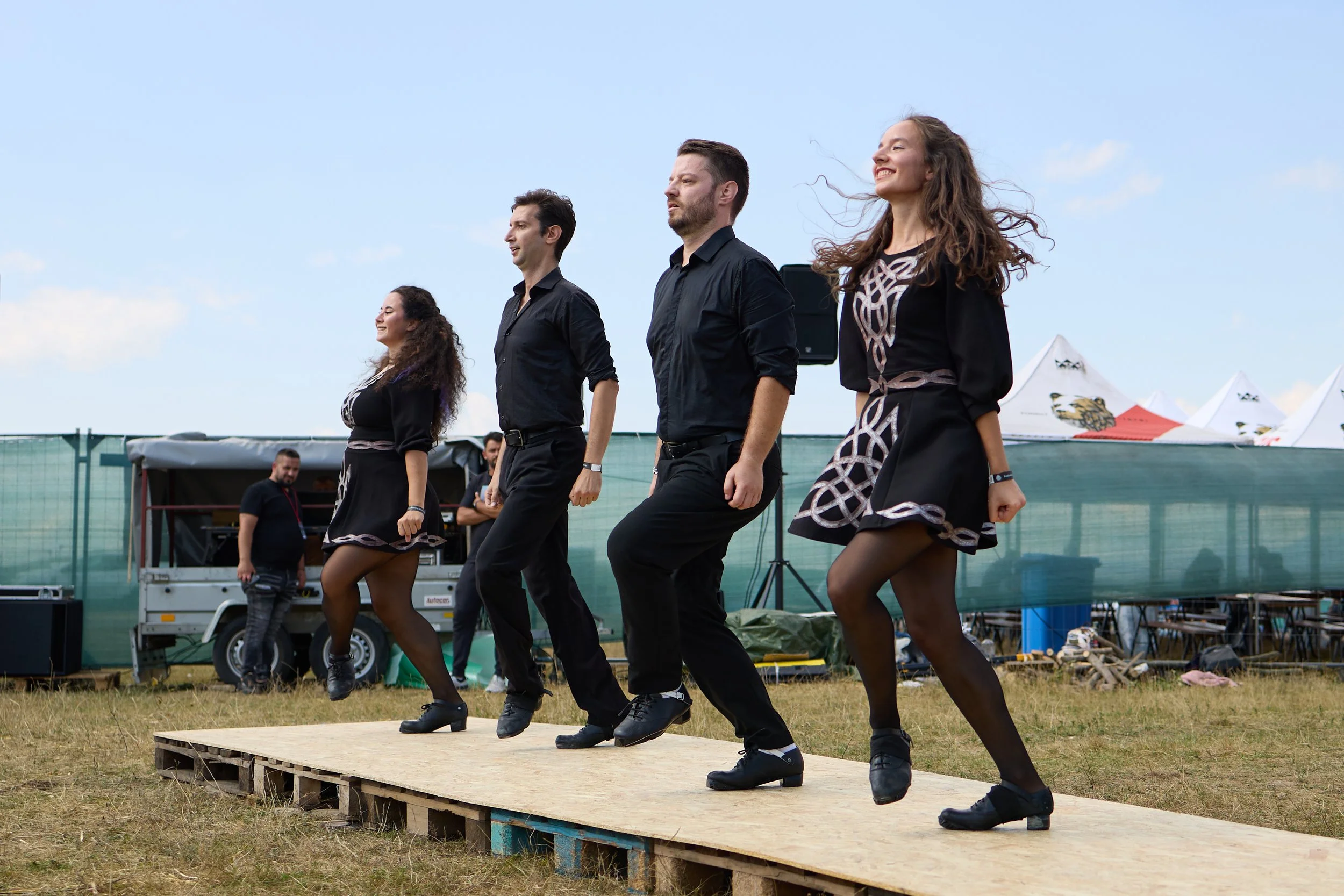Four dancers dressed in black perform a traditional dance on an outdoor wooden stage. They are aligned in a line and smiling, with a clear blue sky above and tents in the background.
