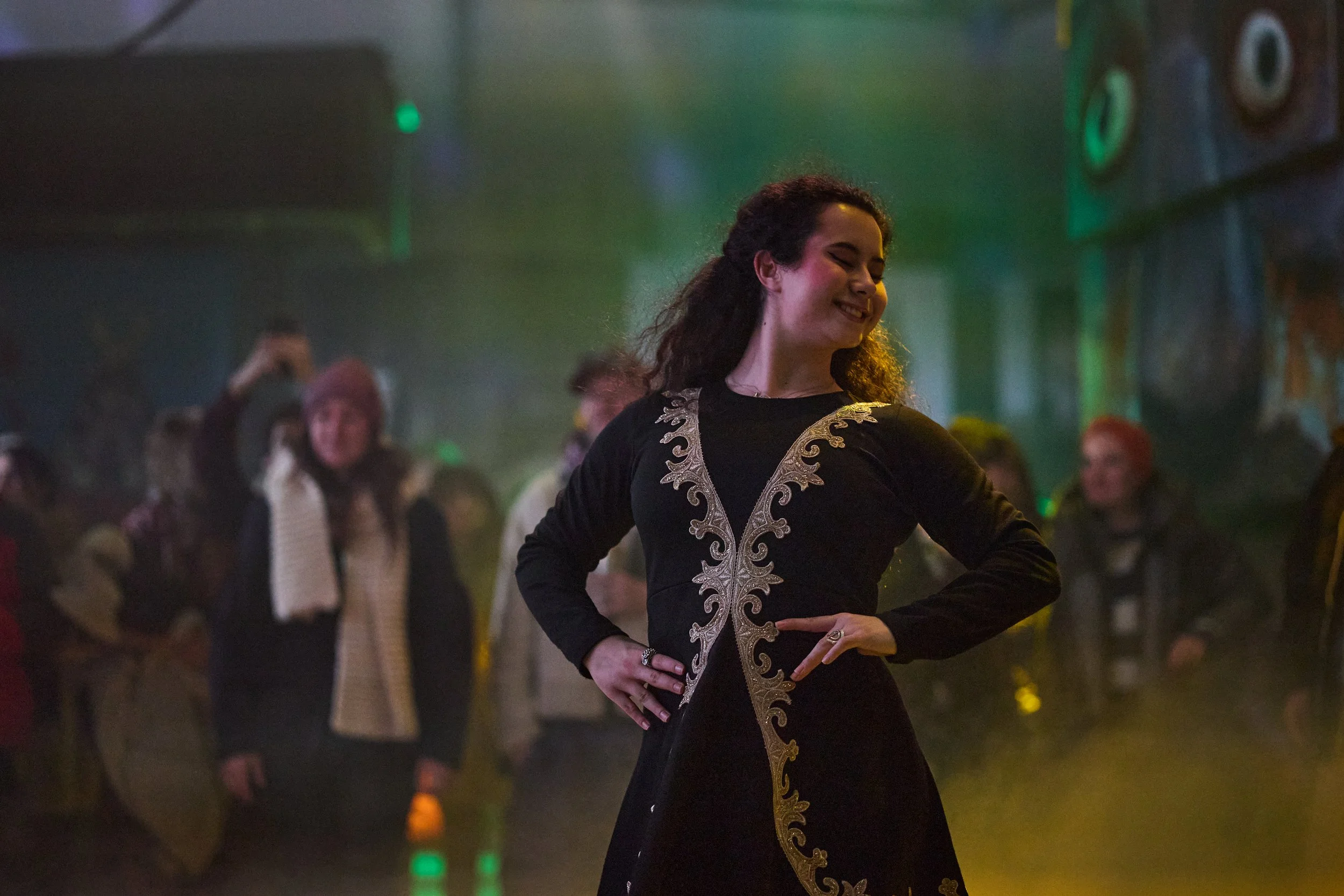 A woman with dark, curly hair smiles as she dances at a social event. She is wearing a black dress with gold embroidery along the front. In the background, several people are also dancing or socializing in a dimly lit room with green and yellow light