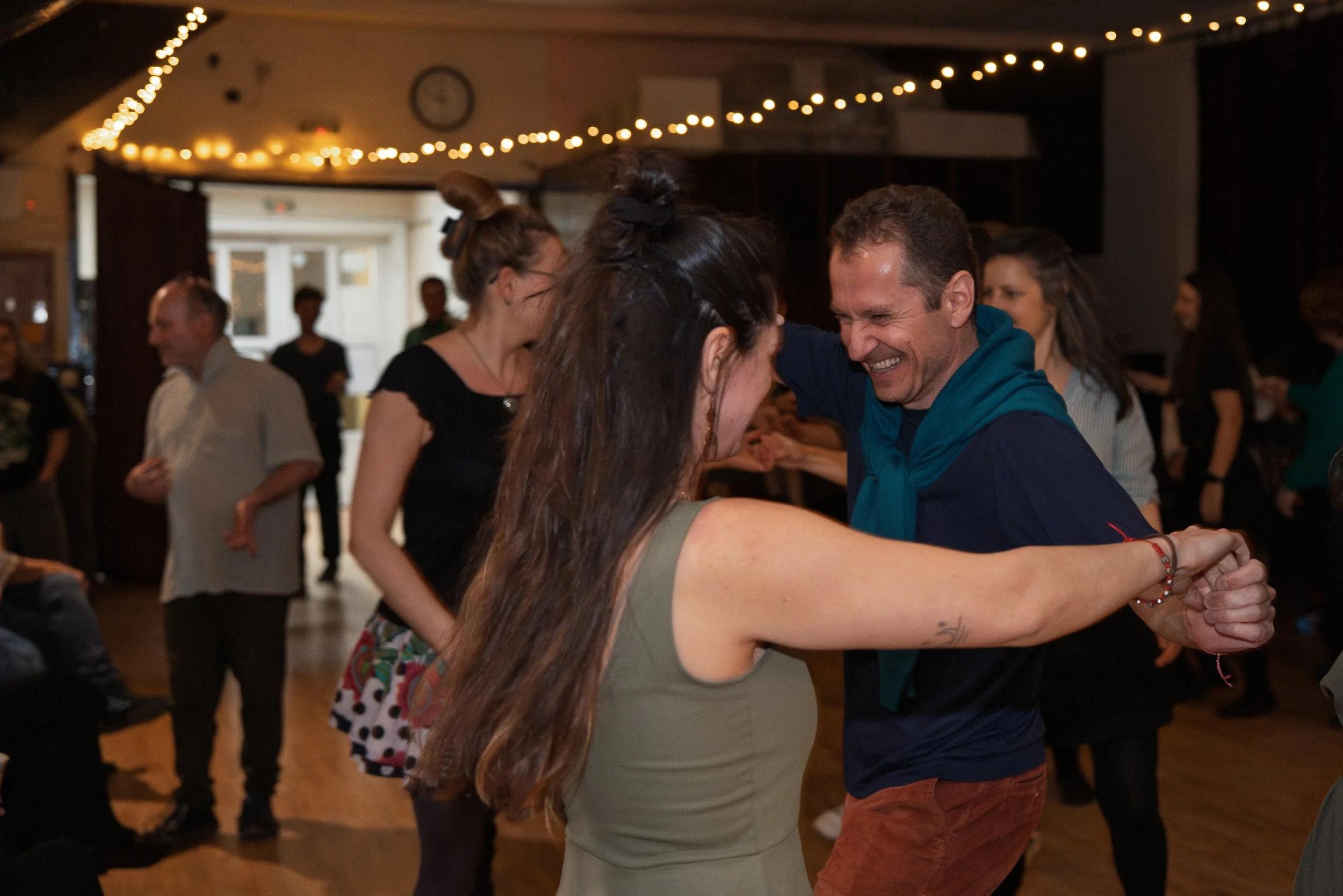 Couple dancing closely in a dimly lit room decorated with string lights, with other people socializing in the background.