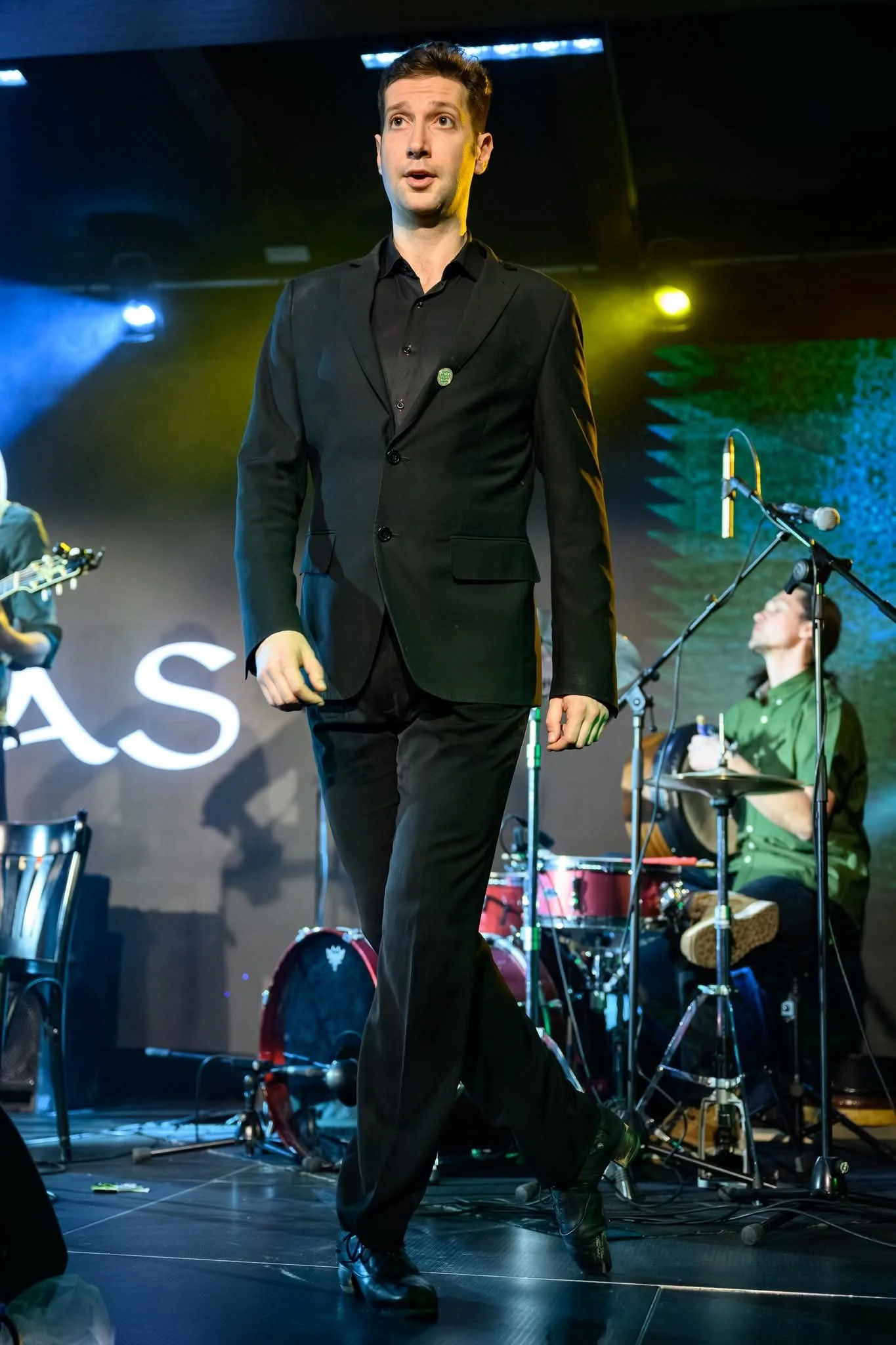 A man in a black suit walking on stage during a concert or event, with musical instruments and other performers visible in the background.