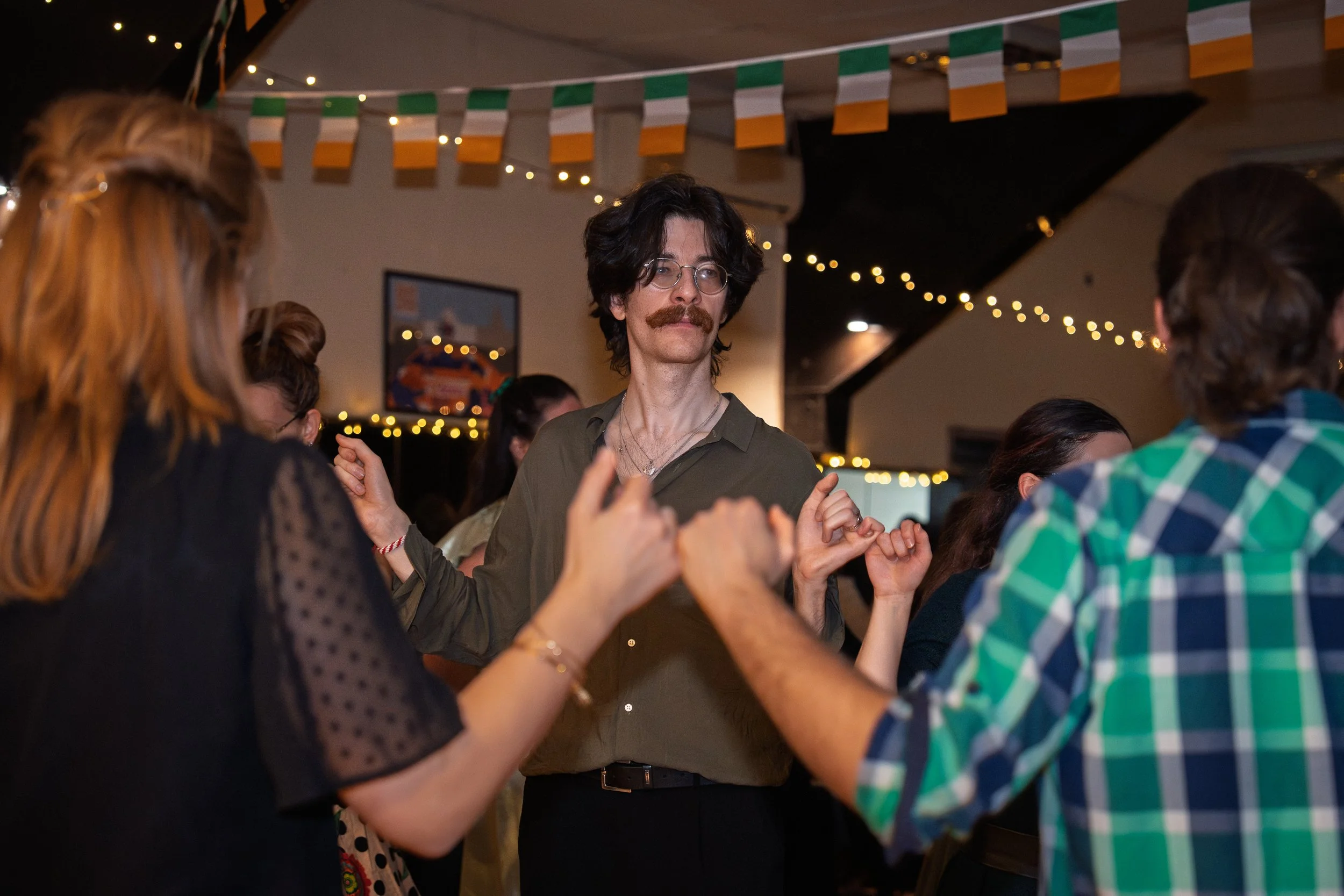 People dancing closely at a celebration or party in a decorated indoor venue with string lights and green, white, and orange banners.