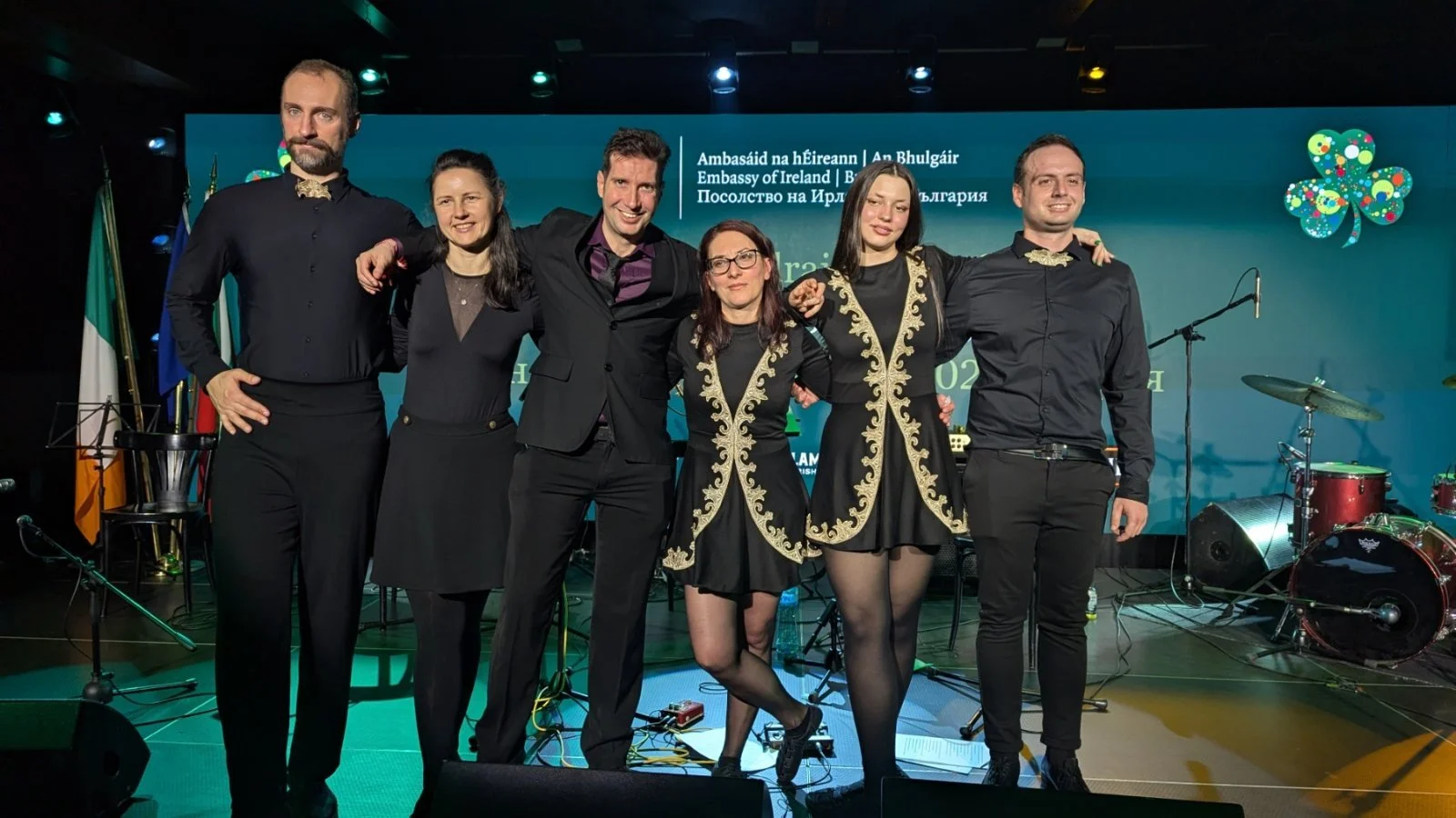 Six people standing on stage with musical instruments in the background, at an event with a blue backdrop displaying Irish and Bulgarian text and the Irish and Bulgarian flags.