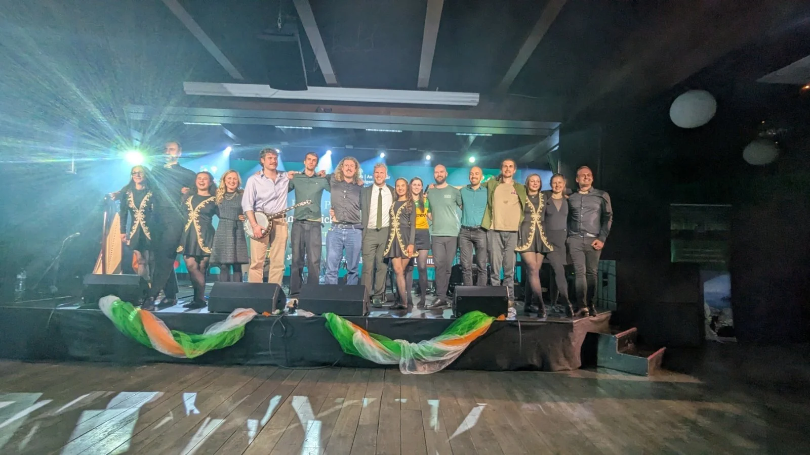 Group of performers and audience members on stage, with some holding musical instruments, in a concert hall with stage lighting and decorations.