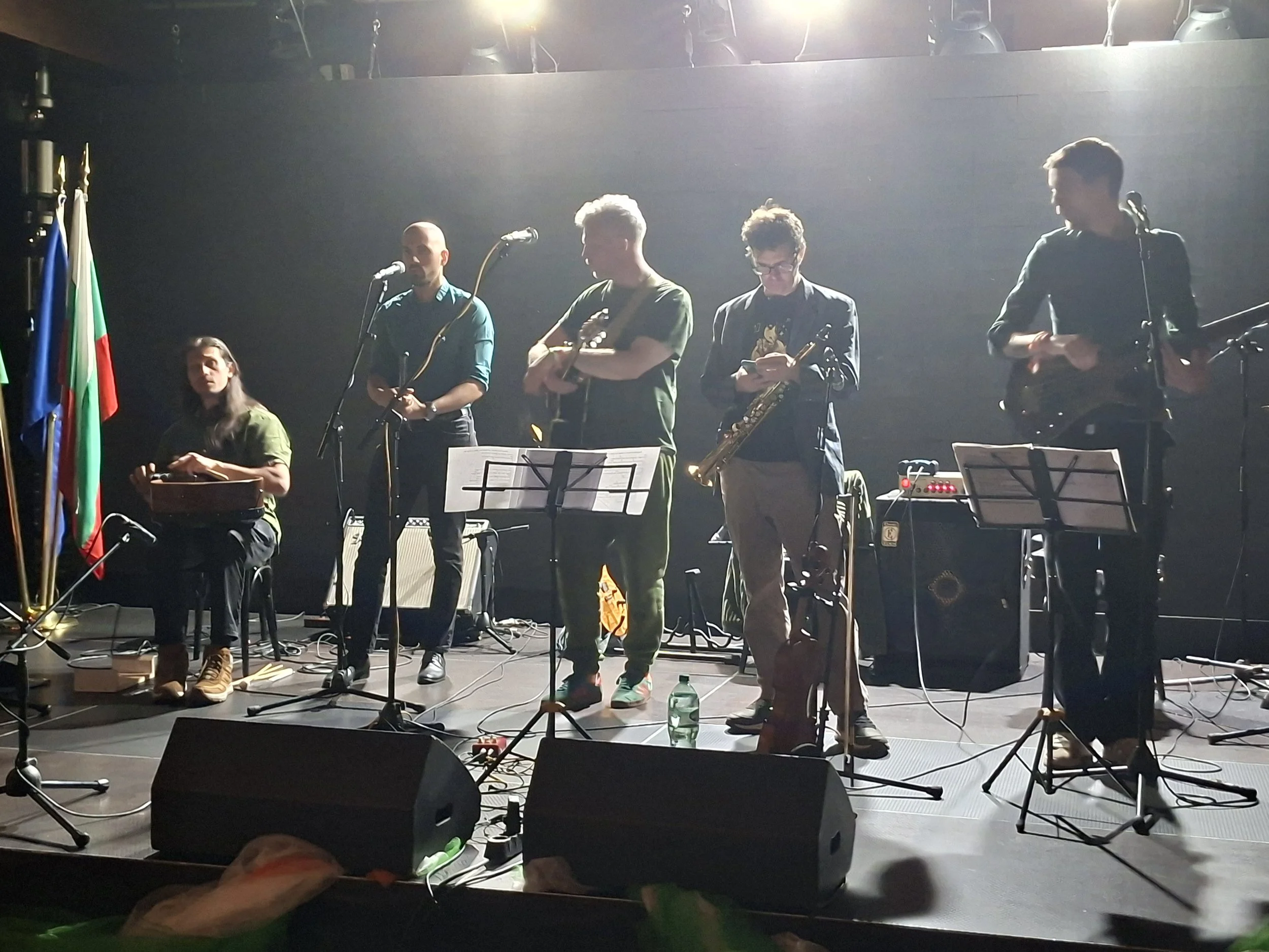 A group of five musicians on stage with musical instruments, including microphones, a violin, and a saxophone. The stage is dimly lit with some stage lighting, and there are flags on the left side of the image. The musicians appear to be preparing to