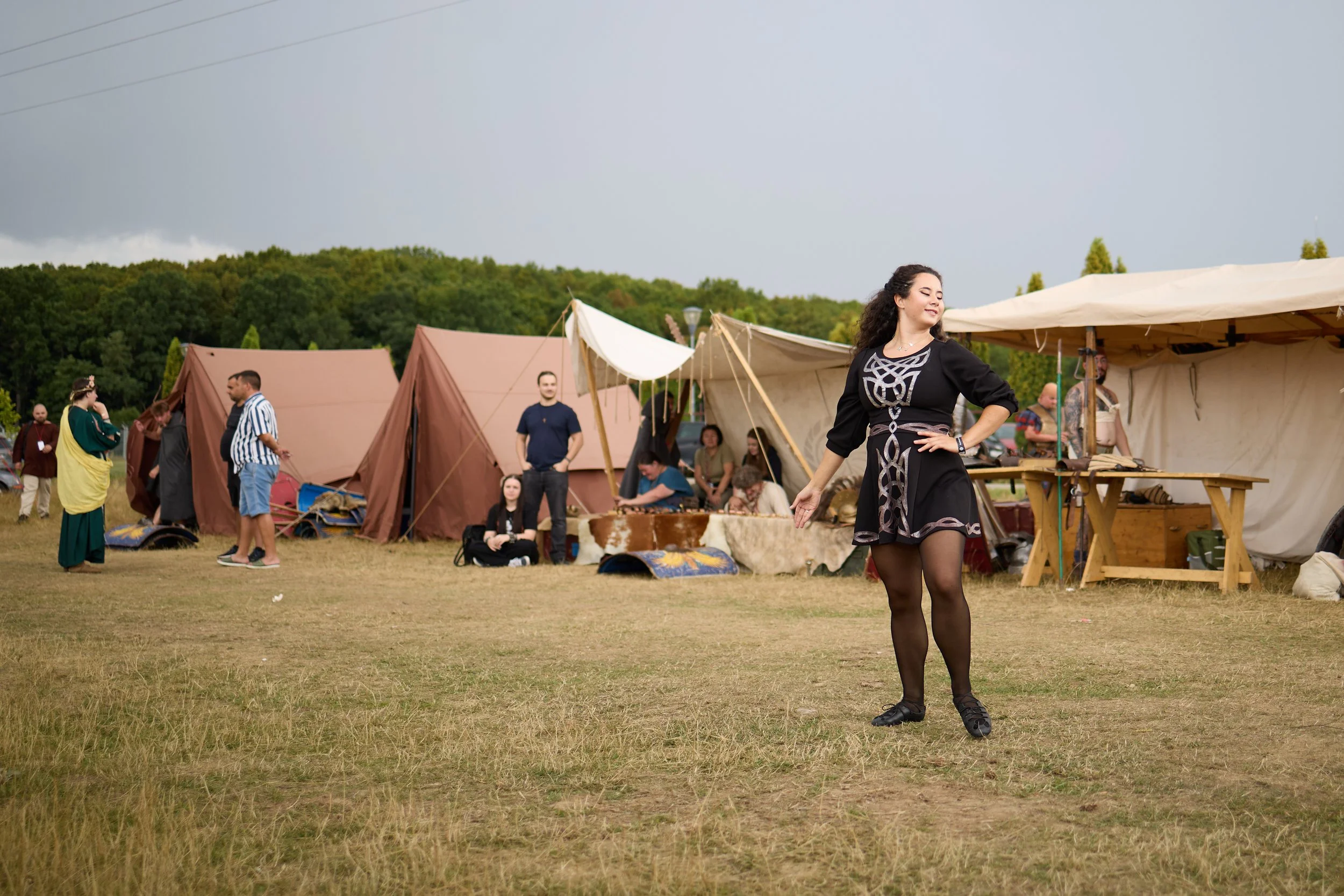 A woman with dark curly hair in medieval costume standing outdoors at a historical reenactment festival with tents and people in the background.