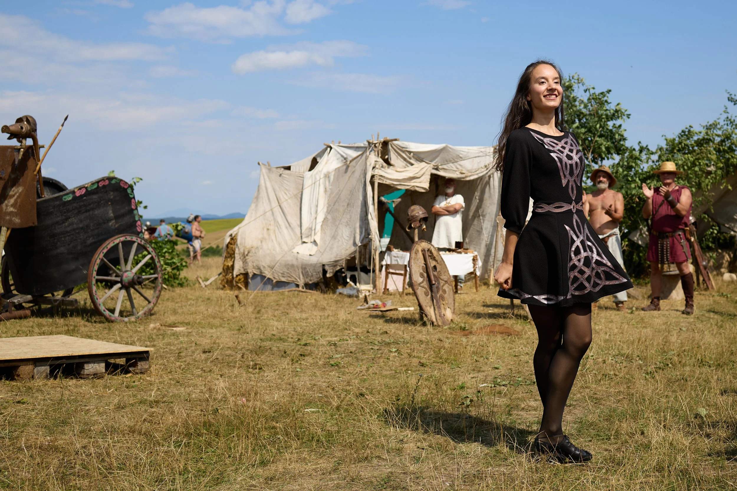 A woman in a black dress with pink designs walking outdoors at a historical reenactment event with tents, people, and a blue sky in the background.