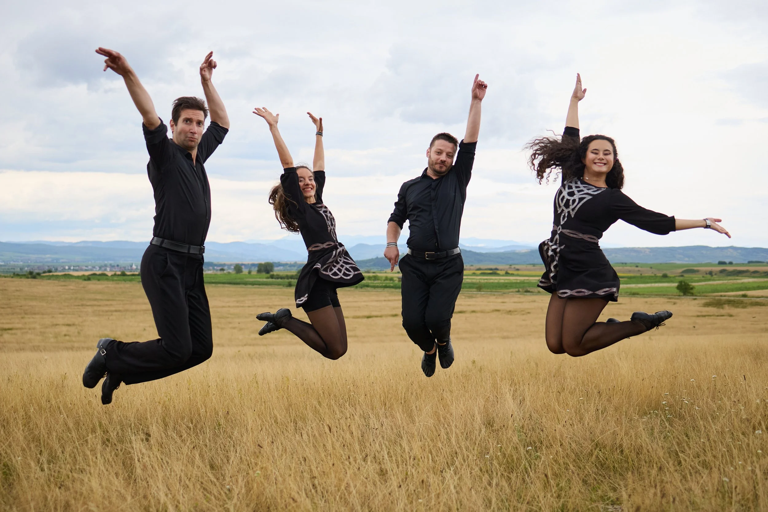 Four people jumping in the air in a field with mountains and cloudy sky in the background. They are smiling and appear happy.