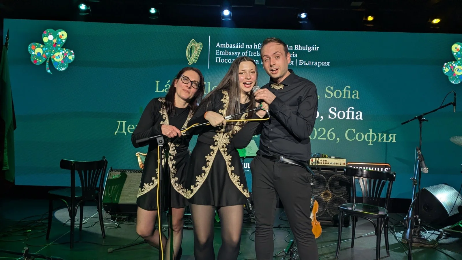 Three people singing and performing on a stage at an event, with a green background displaying text in multiple languages and the Irish and Bulgarian emblems. The two women and one man are dressed in black with gold accents, and they are holding micr