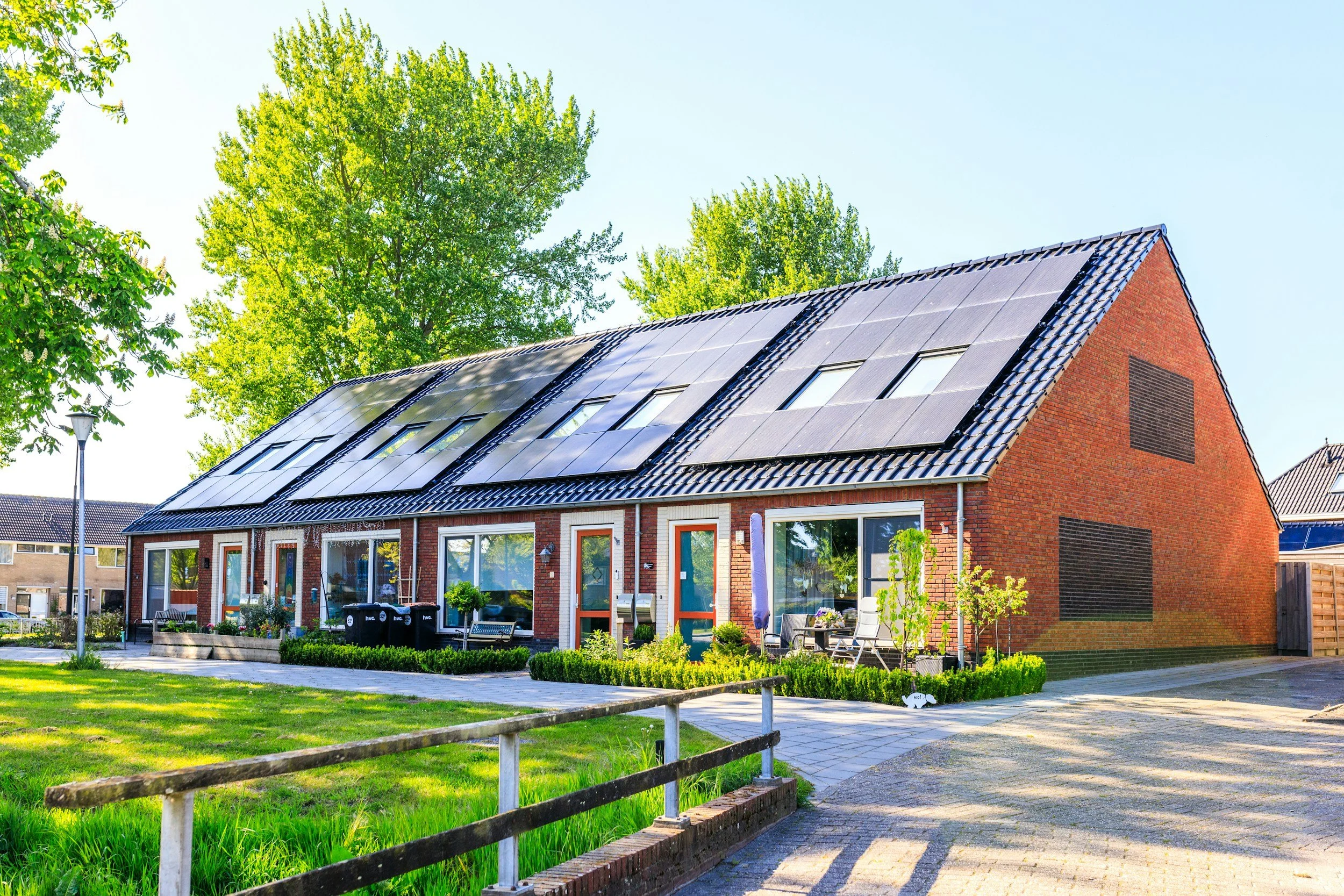 Red brick house with solar panels on the roof, surrounded by green trees and a garden, with a paved walkway and a street lamp nearby.