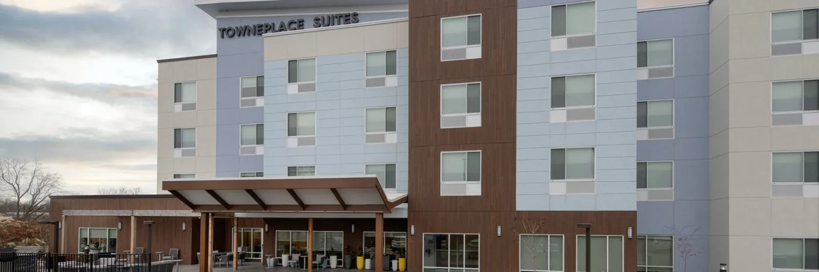 Exterior view of the Townplace Suites hotel showing a modern multi-story building with blue and beige panels, a brown accent section, and an entrance with a covered patio area.
