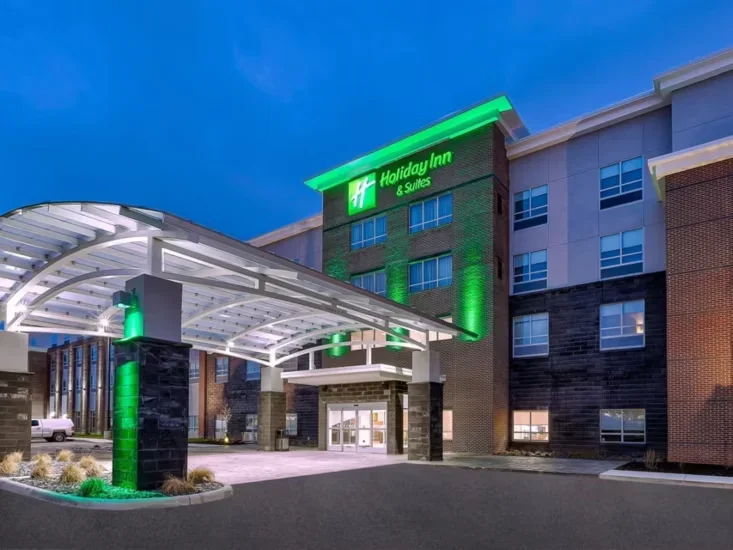 Exterior view of a Holiday Inn & Suites hotel with a green lit sign, entrance canopy, and modern architecture on a clear evening.